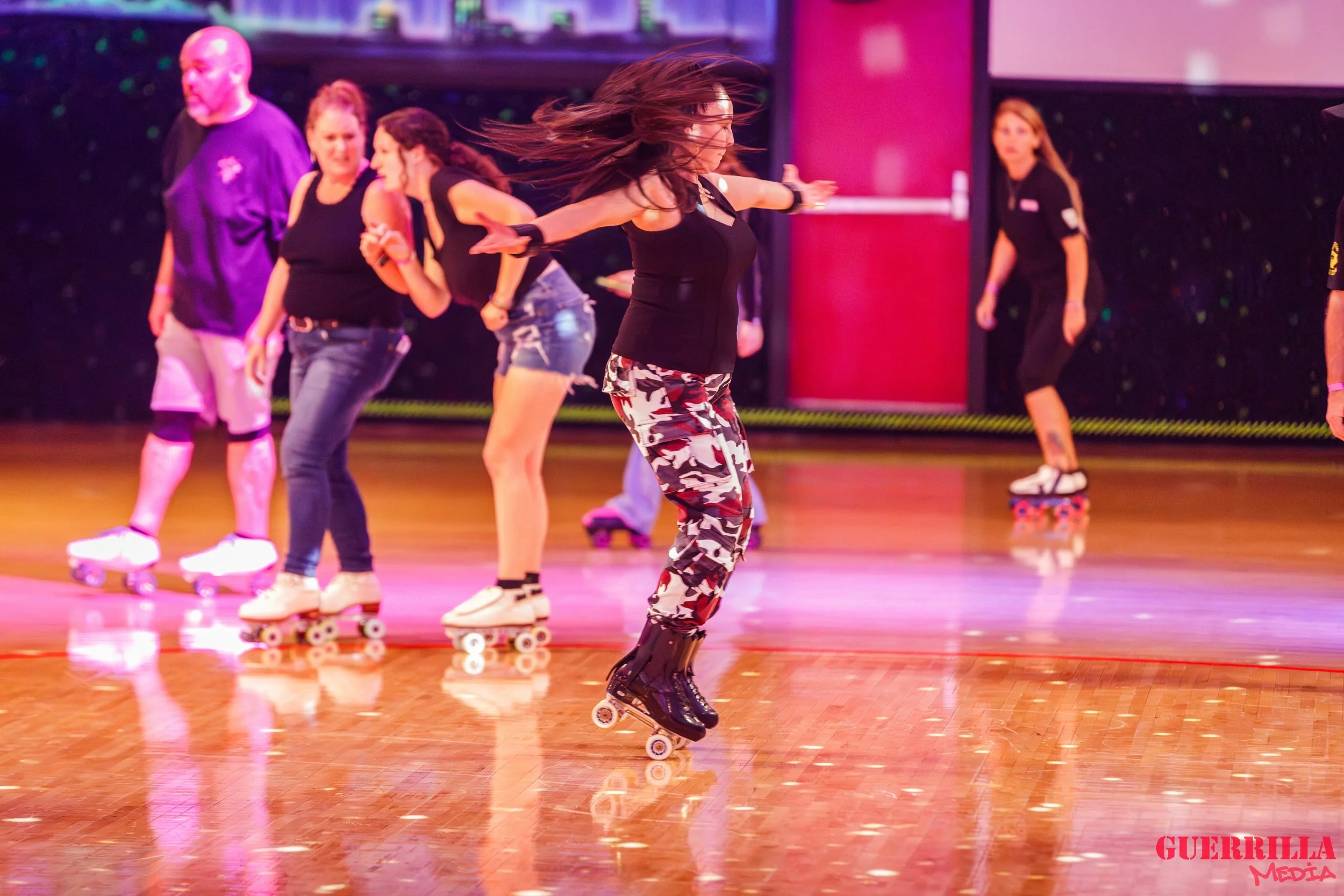 A group of women roller skating indoors on a polished wooden floor, with colorful lighting and some spectators in the background, including a man with a beard.