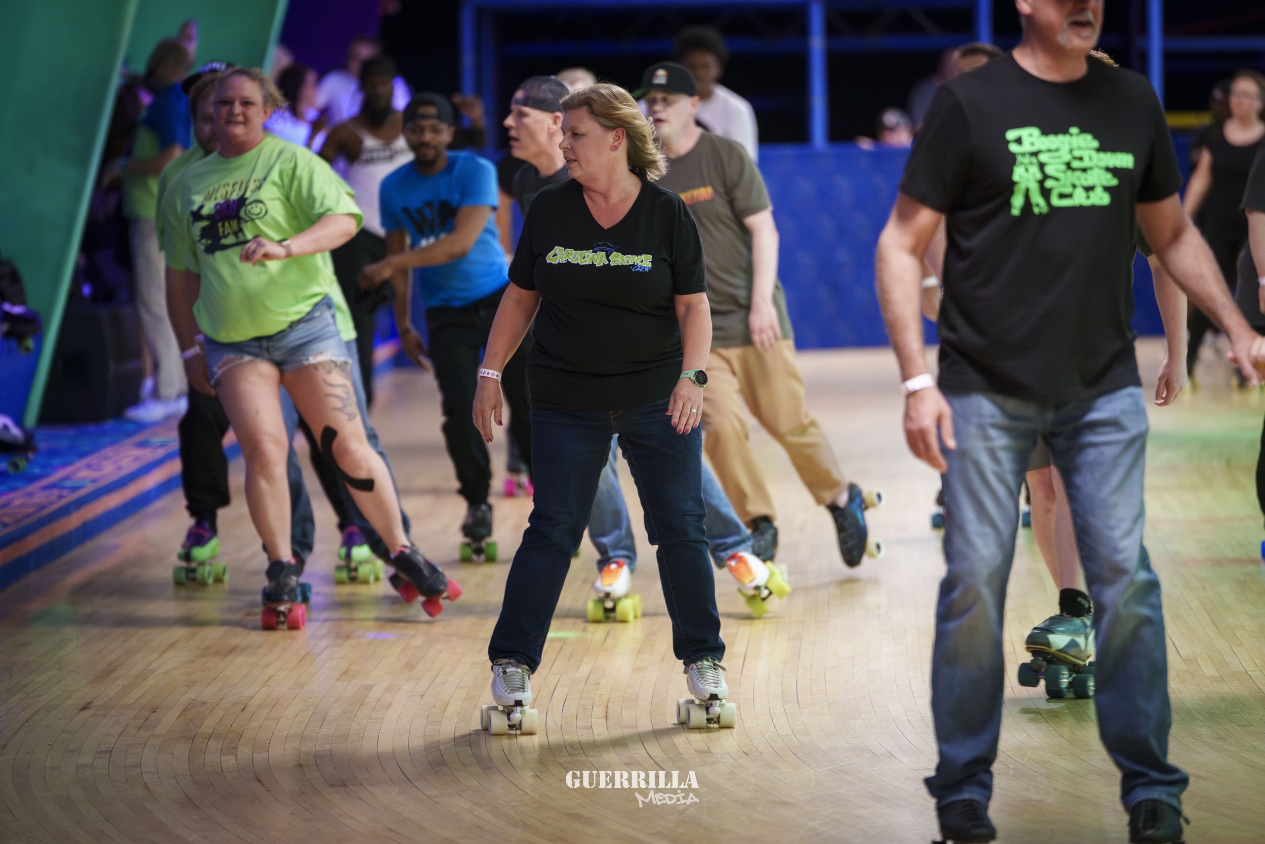 People roller skating indoors, some wearing casual clothes, others in event T-shirts, with colorful lighting and a wooden floor.