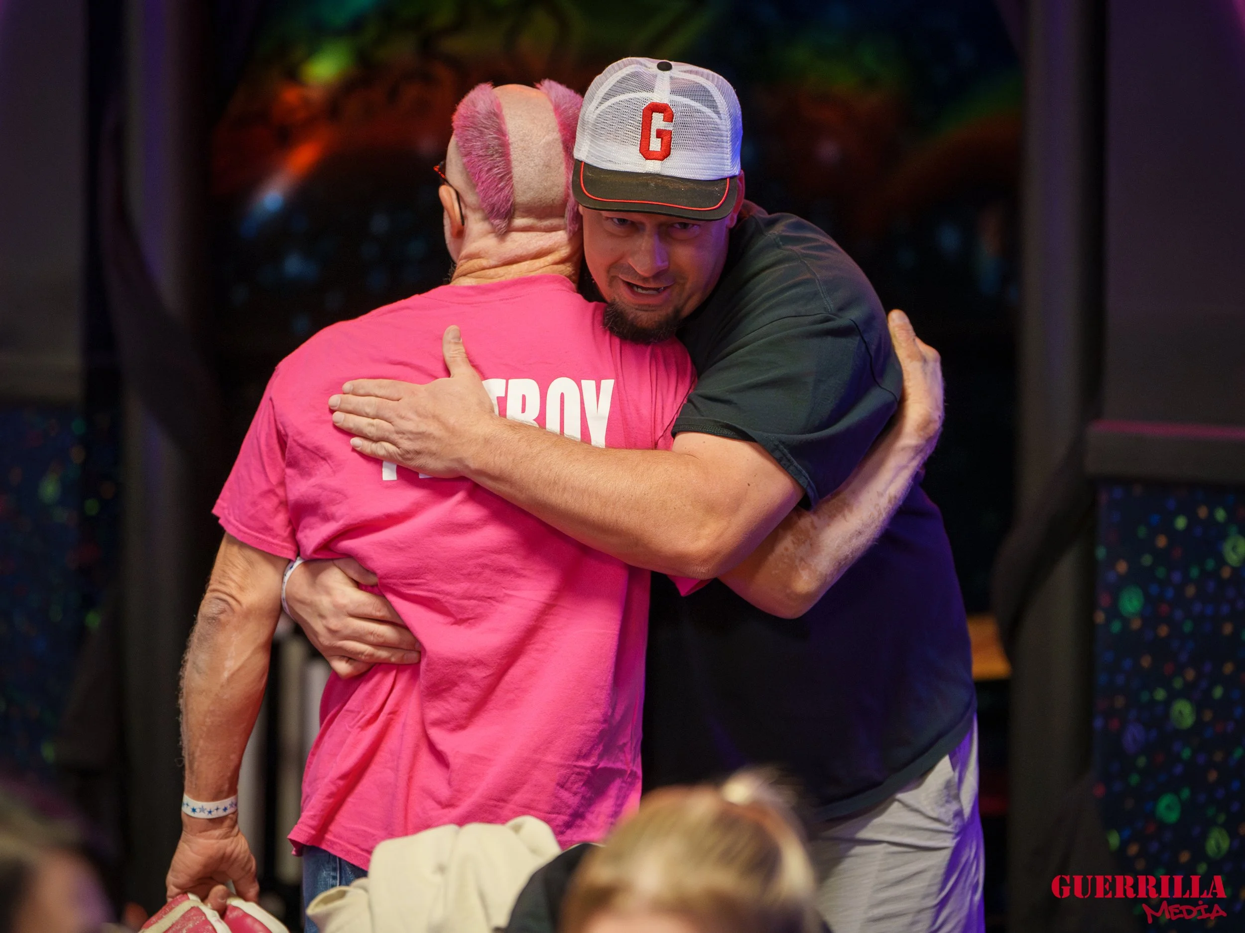 Two men hugging each other warmly on a stage or in a public setting, one wearing a pink shirt and cap, the other in a black shirt and a white cap with a red letter 'G' on it.