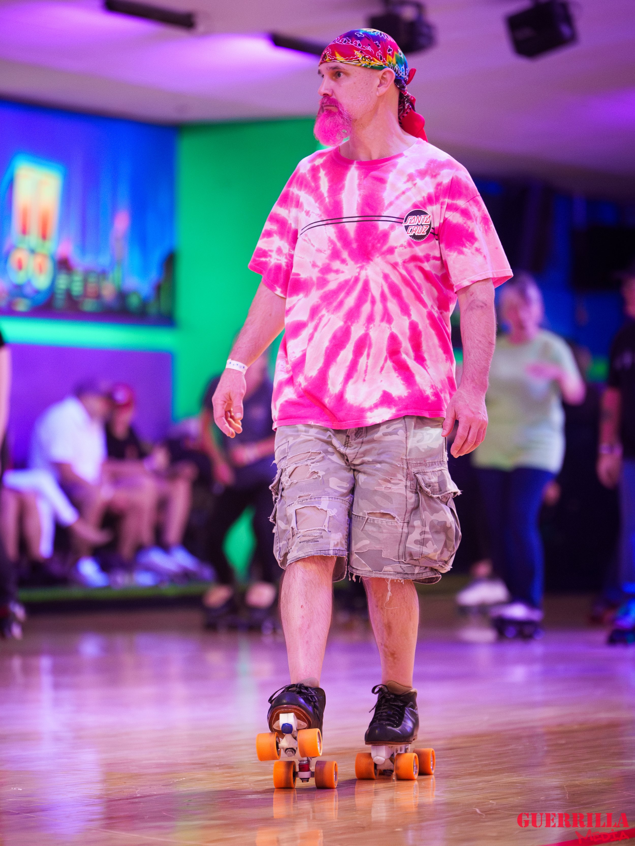 A man with a pink tie-dye T-shirt, camouflage cargo shorts, black roller skates, and a colorful bandana skating on a roller rink with neon lights and other skaters in the background.
