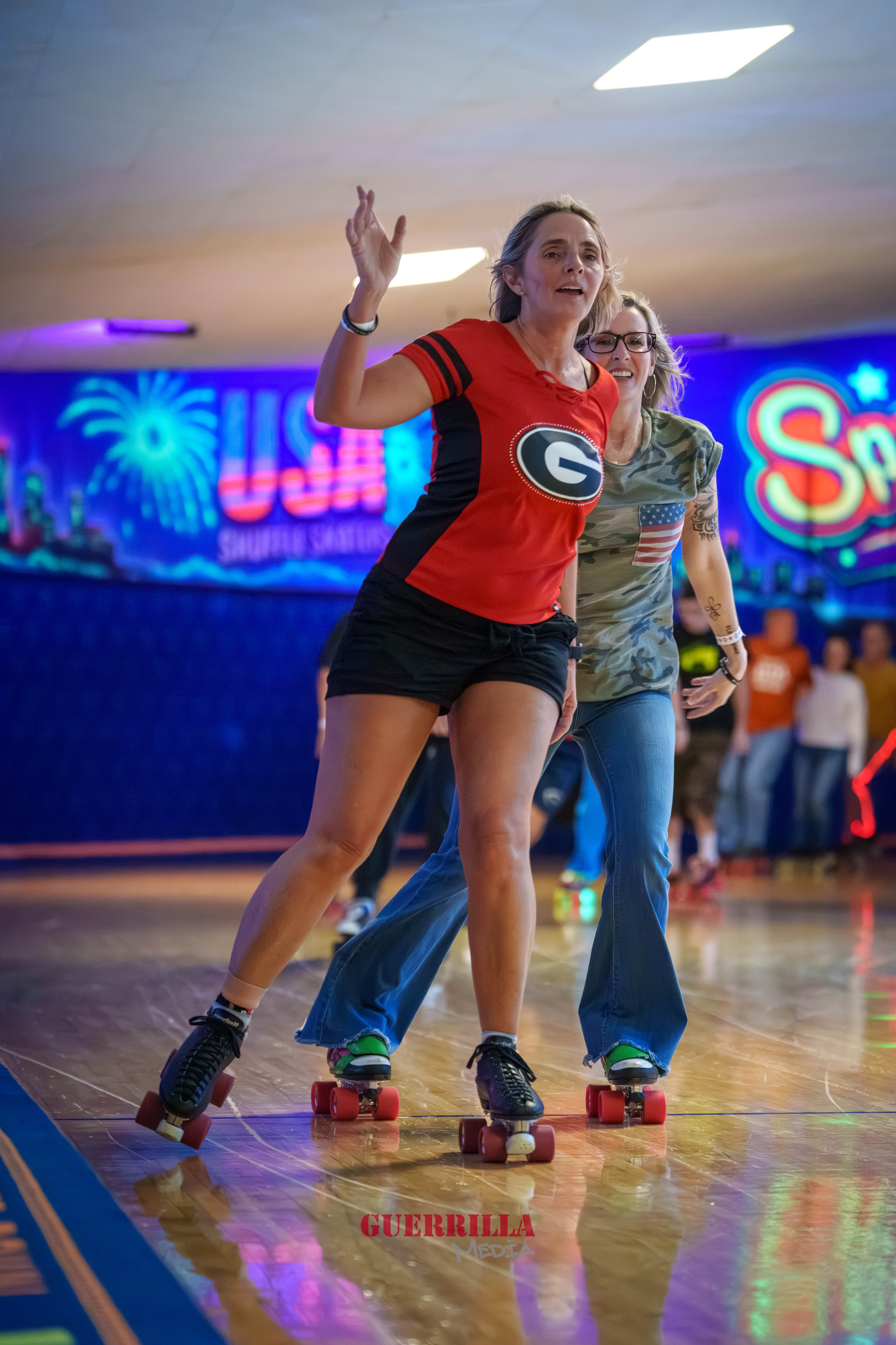 Two women roller skating indoors at a roller skating rink with neon signs in the background. One woman is wearing a red sports jersey with a 'G' logo, black shorts, and roller skates. The other woman is wearing glasses, a camouflage shirt with an Ame