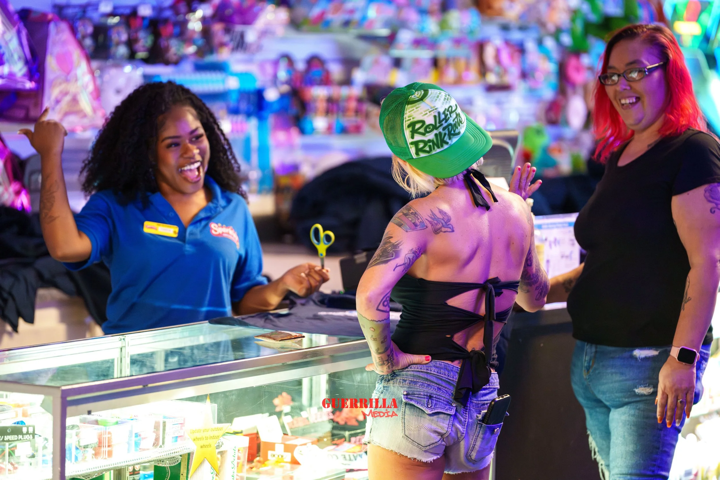Three women are at a colorful arcade or amusement park. One woman, in a blue shirt, is smiling and holding a small pair of scissors, while the other two women are engaged in conversation, one wearing a black top and ripped jeans with tattoos and a gr