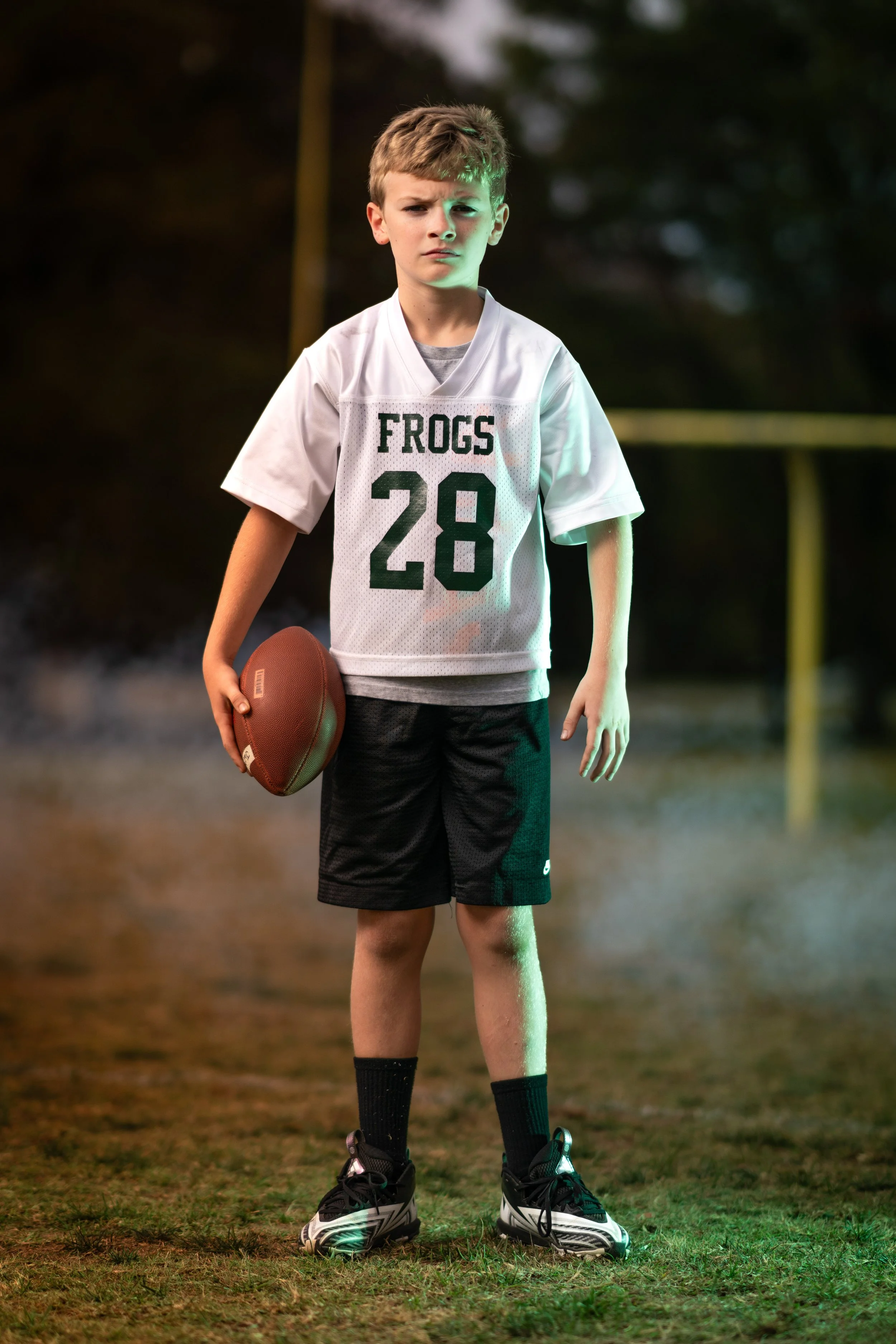 A young boy in a sports uniform holding a football on a grassy field during evening or night time.