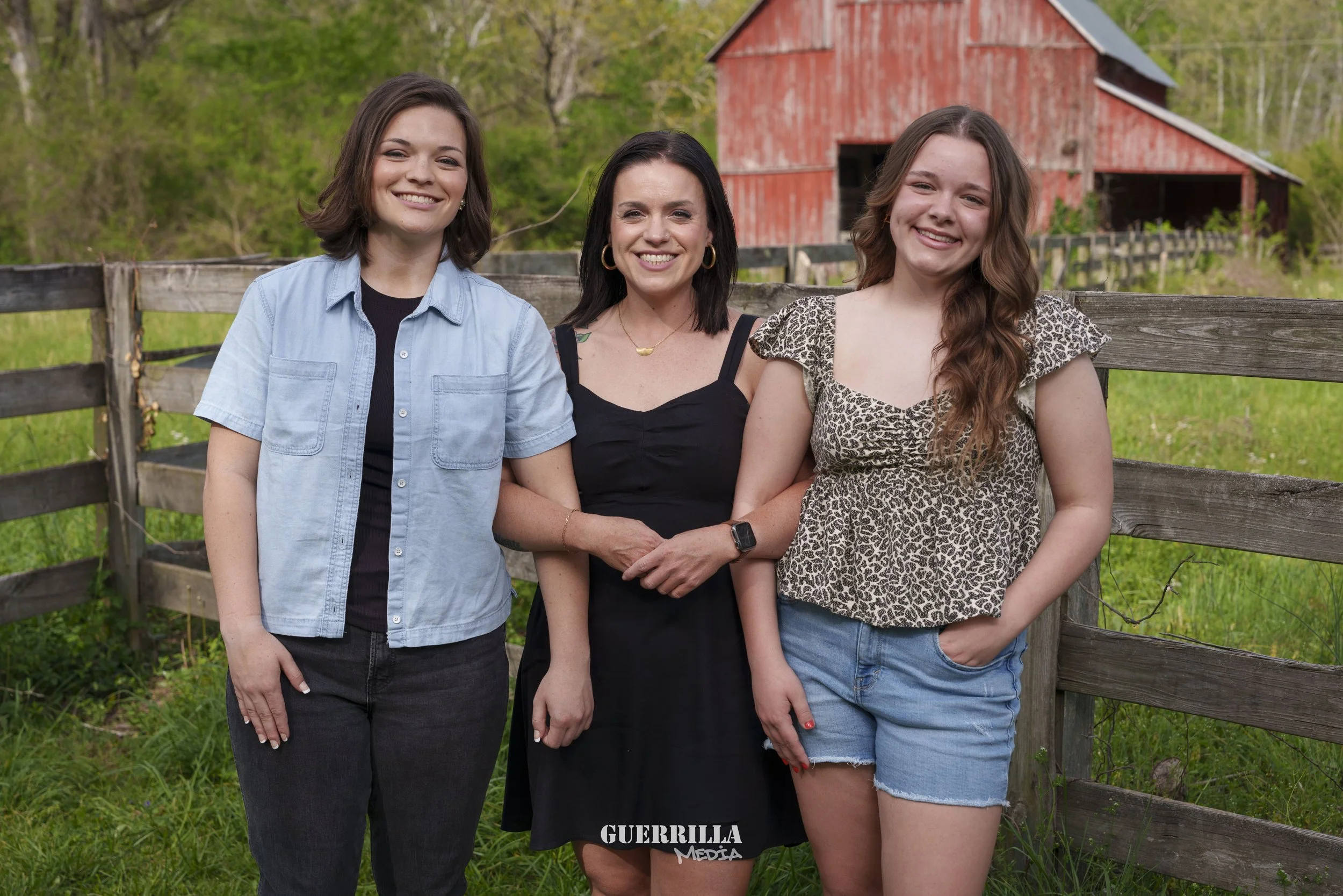 Three women standing outdoors in front of a wooden fence, with a red barn and green trees in the background. They are smiling and posing for the photo.