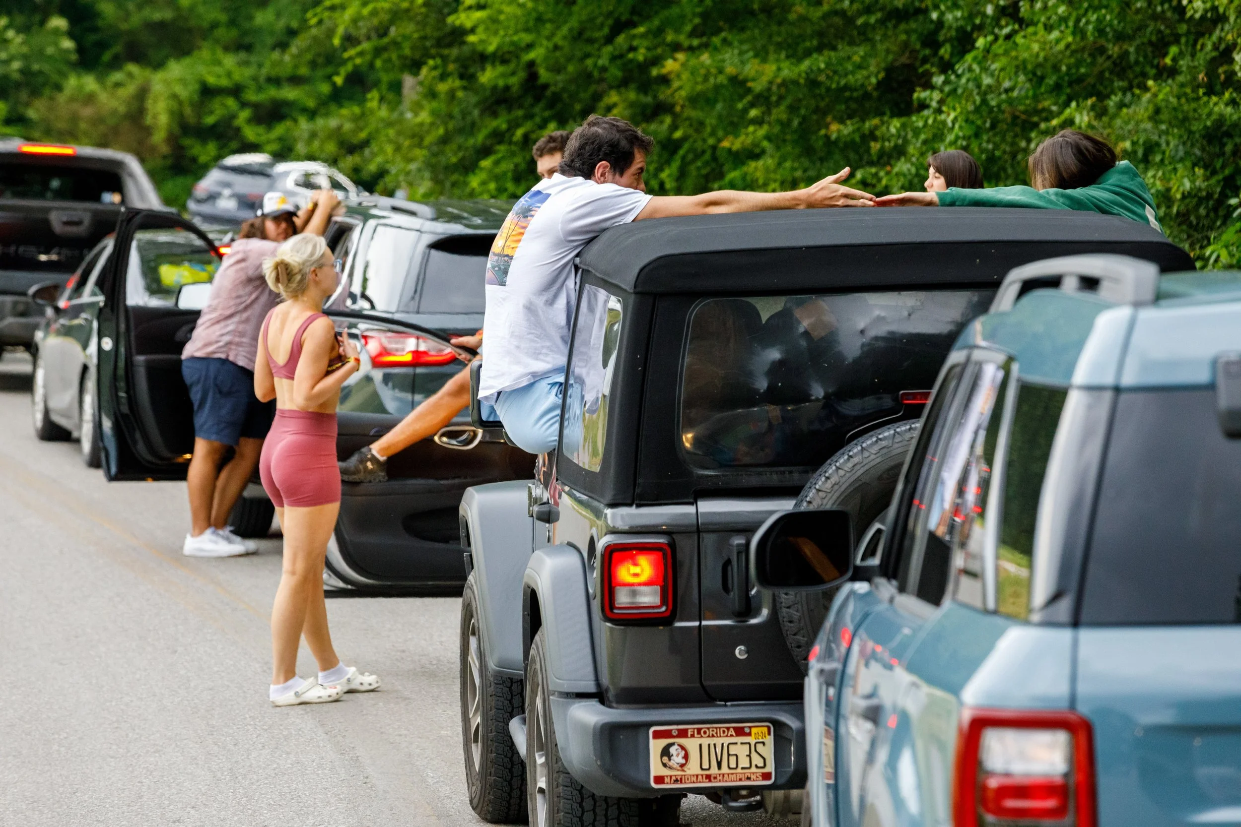 Multiple cars parked on the side of a road with people interacting outside and on top of the vehicles, engaging in conversations and stretching.