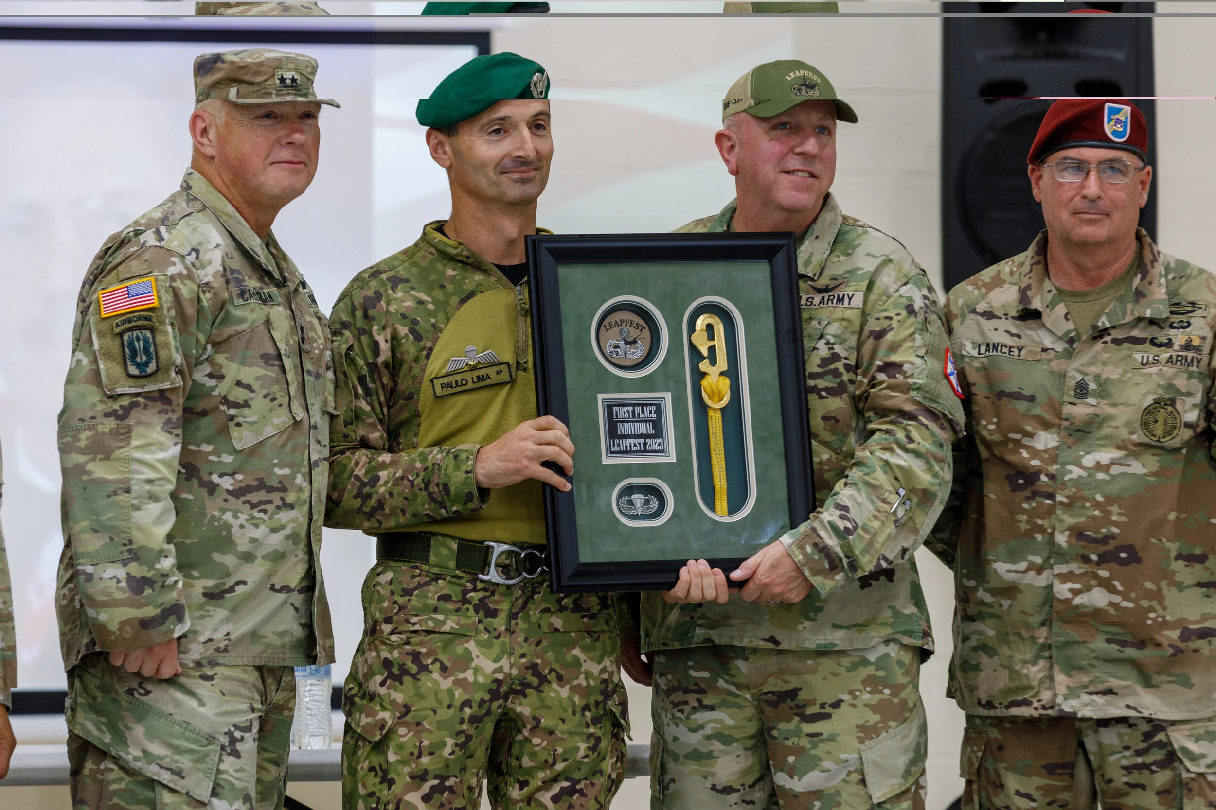 Four military personnel in uniform standing together, holding a framed award or plaque with patches and memorabilia inside, during a ceremony.