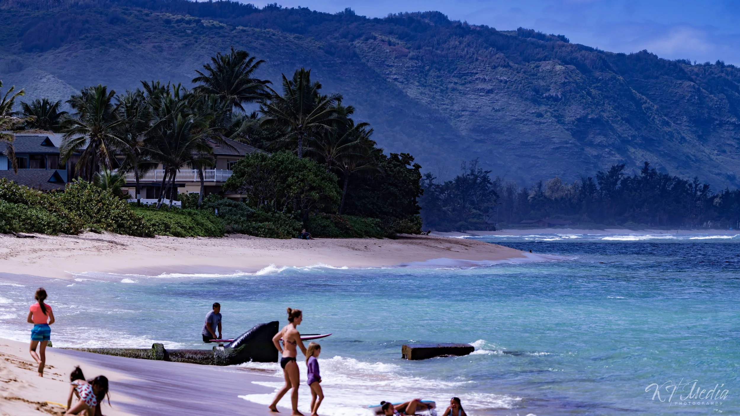 A tropical beach scene with children and adults playing near the shoreline, palm trees, beachfront houses, lush greenery, and mountains in the background