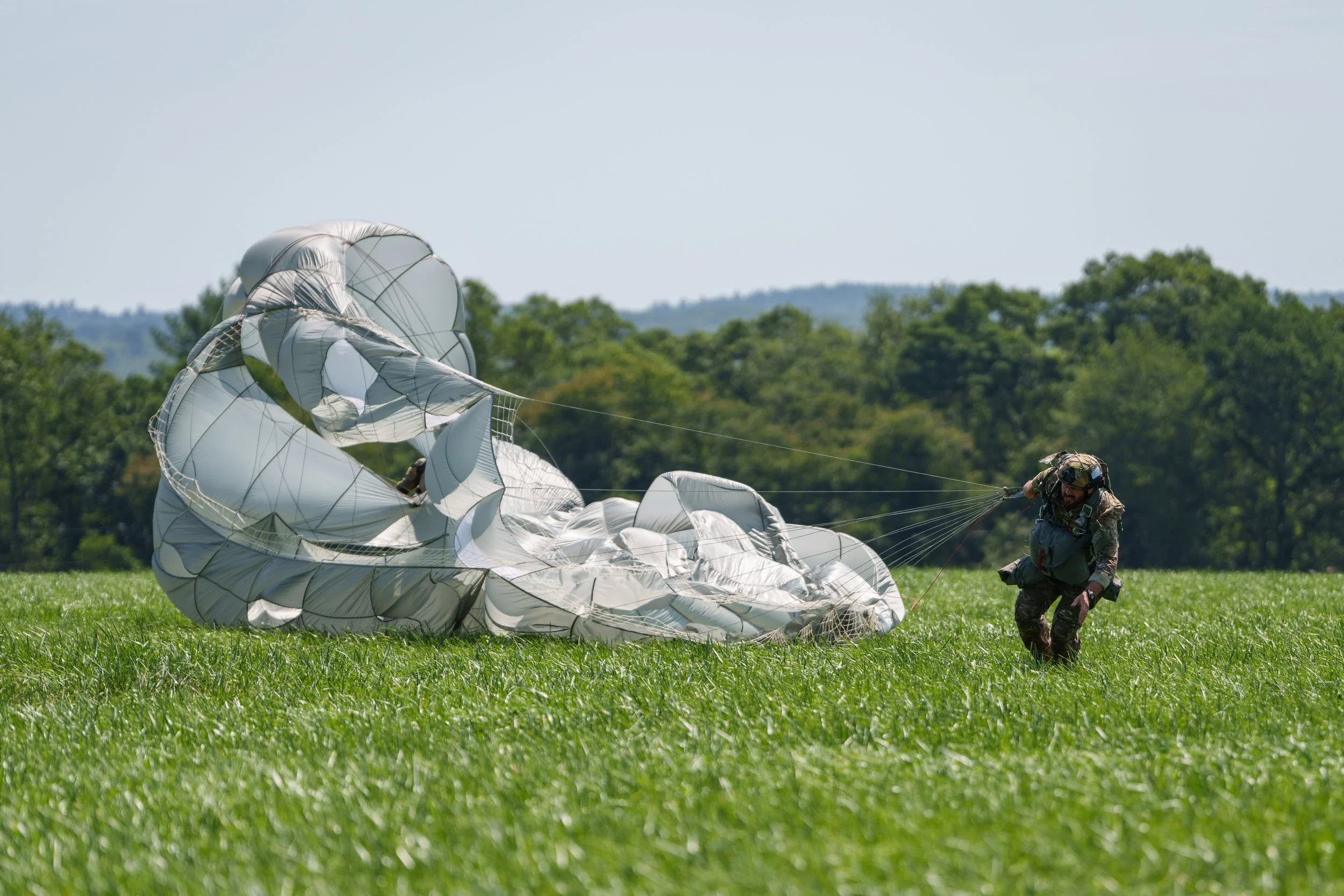 A person in military uniform kneeling on green grass, holding on to a deflated silver parachute with trees and a blue sky in the background.