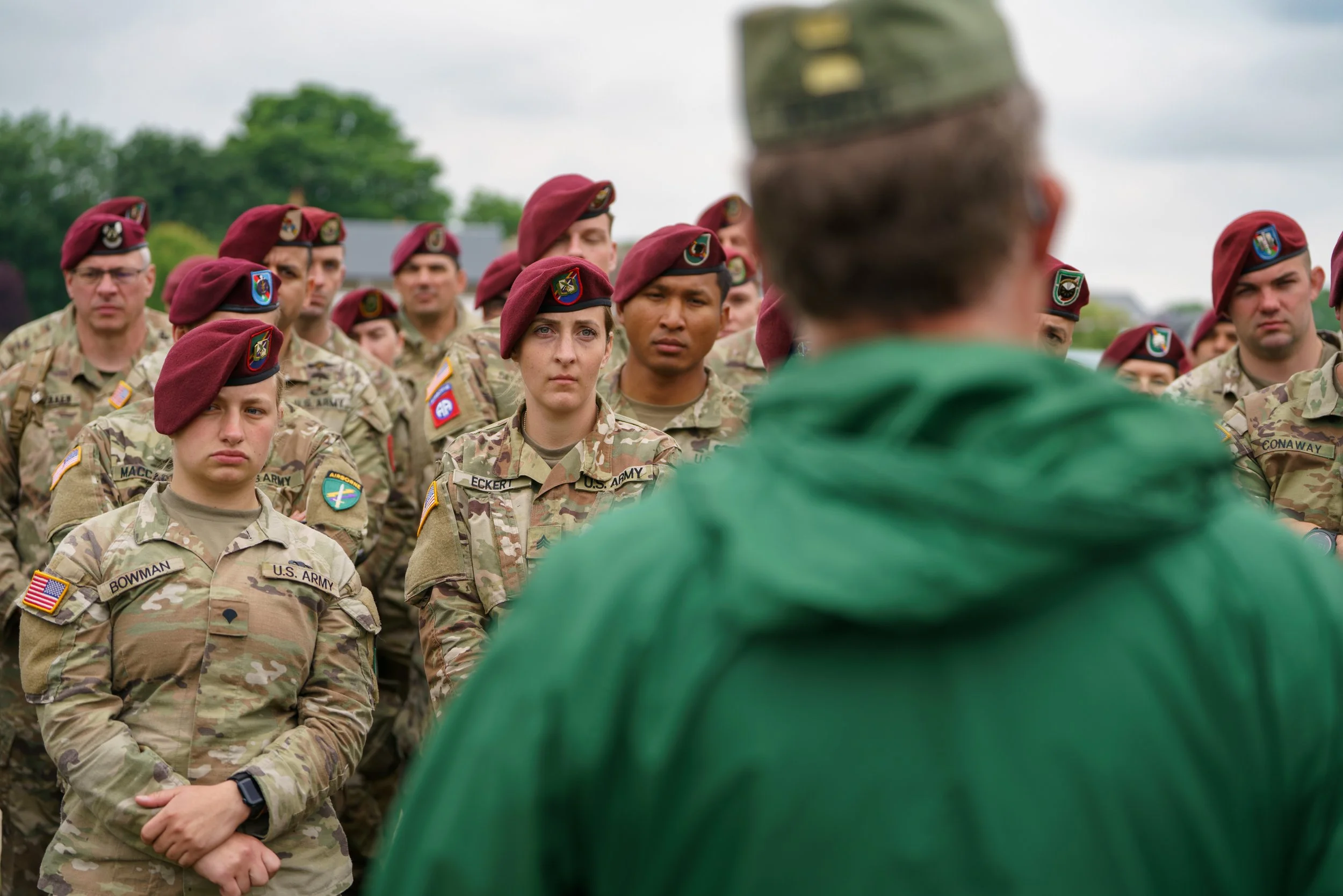 U.S. Army soldiers in camouflage uniforms and maroon berets listening to a speaker in a green jacket outdoors.