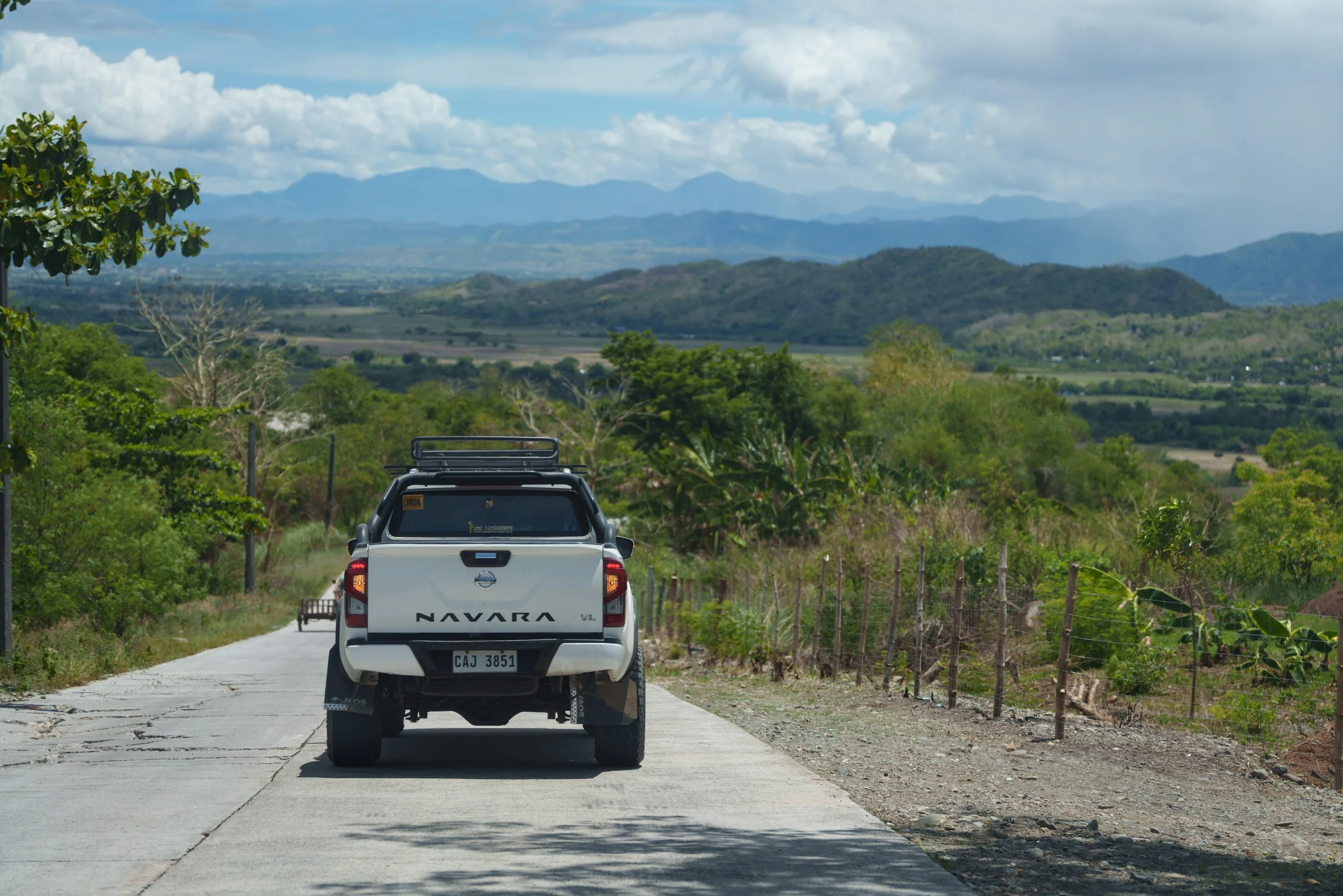 A white pickup truck with 'Navara' written on the back, driving on a rural road surrounded by green trees and shrubs, with mountains and a cloudy sky in the background.