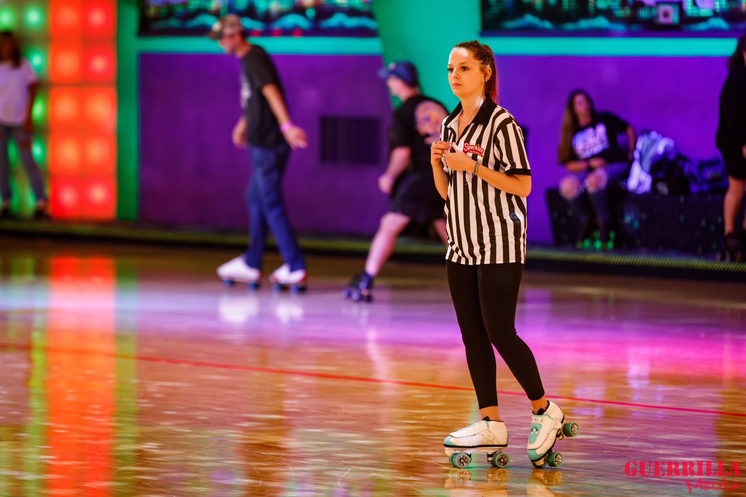 A young woman with red hair in a ponytail skating on a roller rink, wearing black leggings, white roller skates, and a black and white striped referee shirt. In the background, other skaters and spectators are visible with colorful lighting and walls