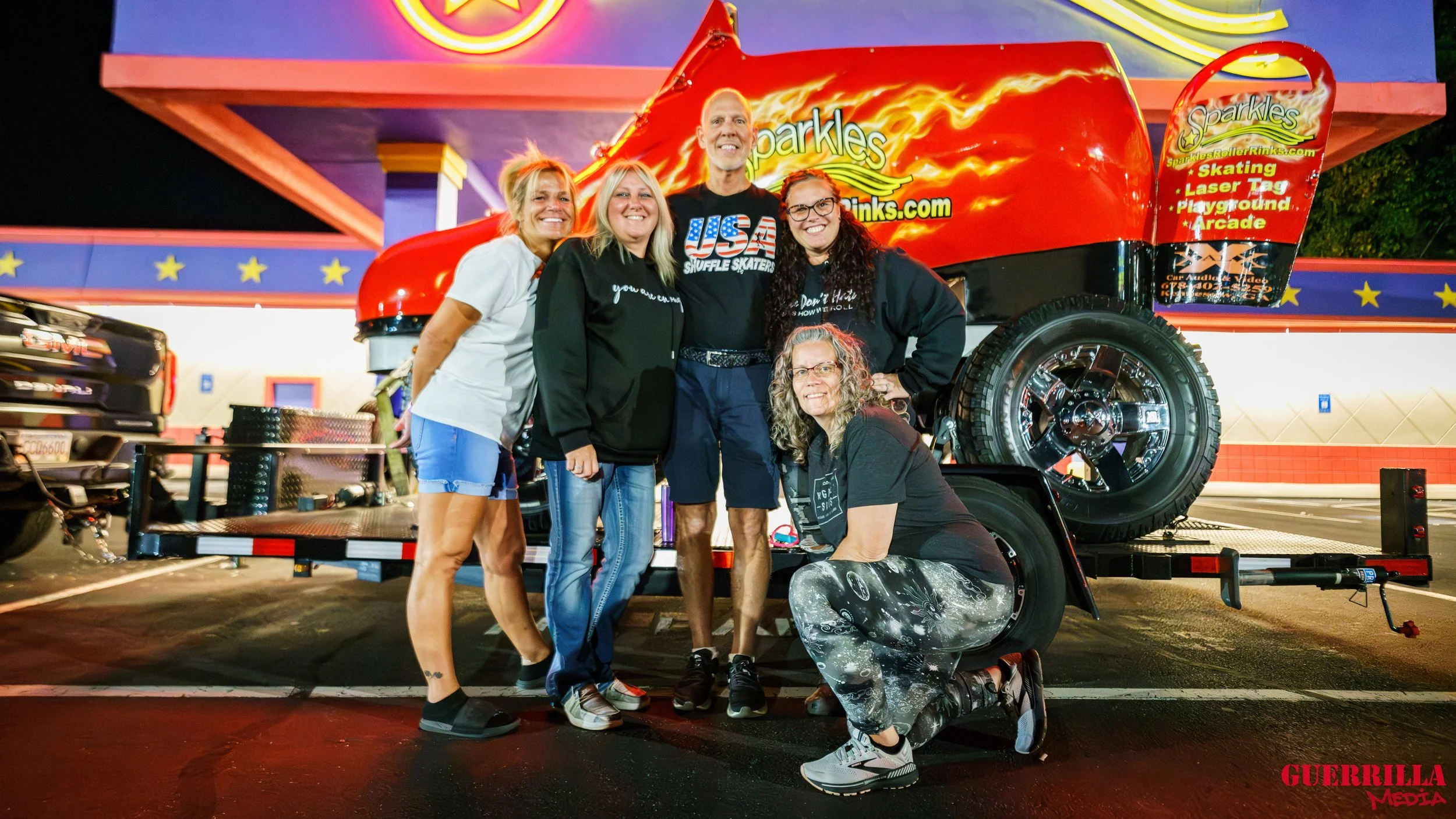 Group of five people smiling and posing in front of a large red amusement ride shaped like a fire truck at night. The background shows a brightly colored amusement area with stars on the walls, featuring the logo 'Sparkles Rollerskating & Rinks'. The