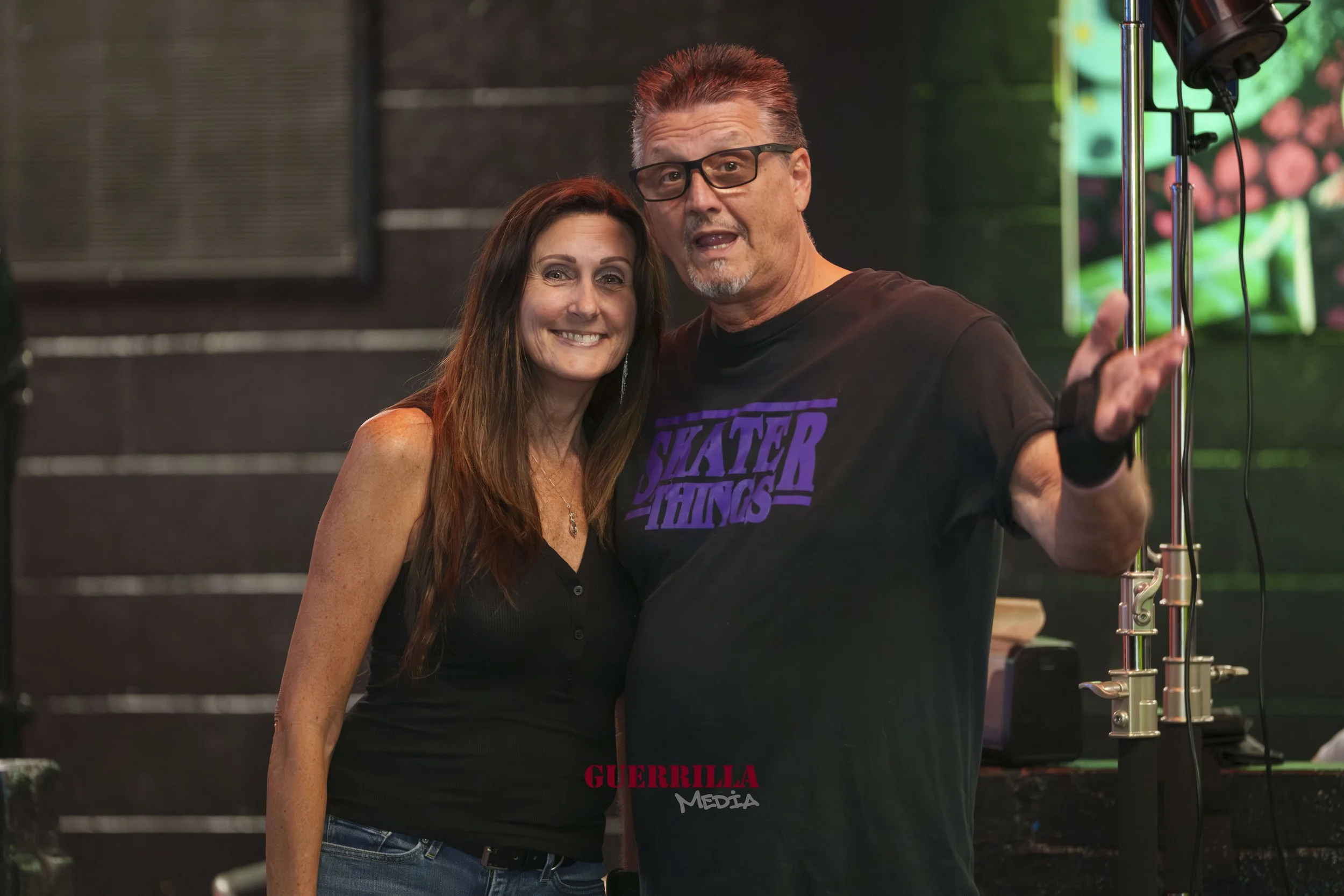 A woman and a man posing together in a dimly lit venue. The woman has long brown hair and is smiling, wearing a black sleeveless top. The man has short spiky hair, glasses, and is wearing a black T-shirt with purple text that reads "Skater Hatings." 