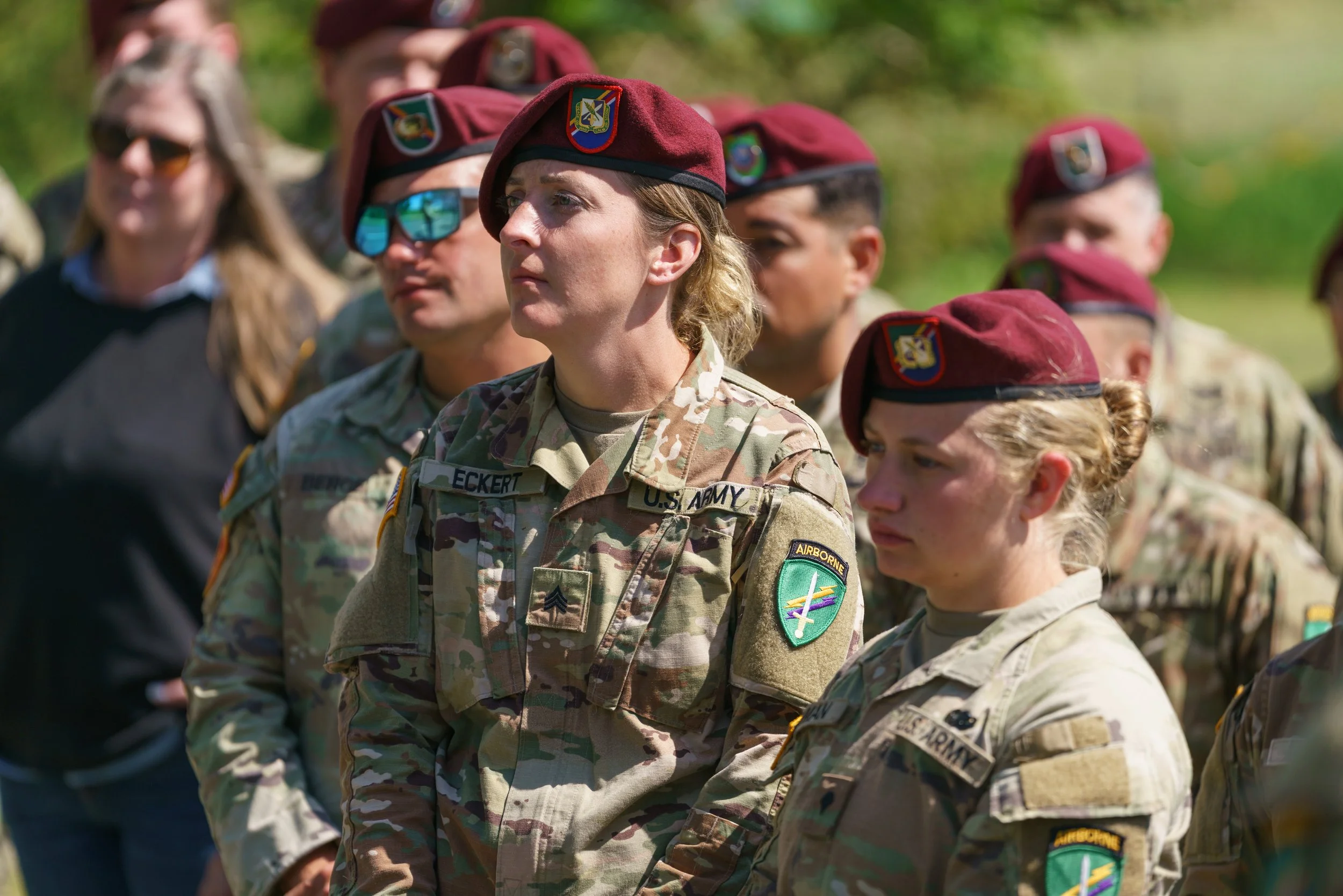 Group of female soldiers in camouflage uniforms and maroon berets standing outdoors, listening attentively.