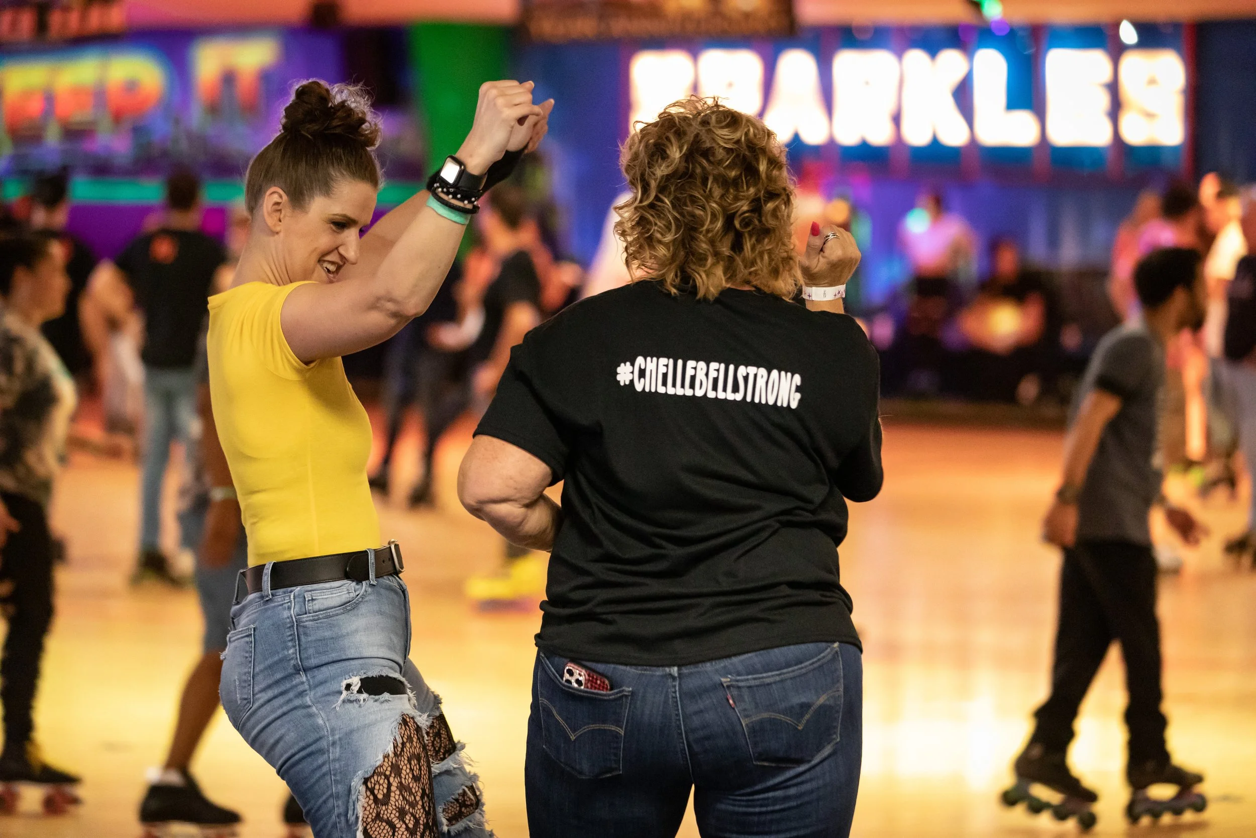 Two women roller skating at an indoor roller rink, with one wearing a yellow shirt and ripped jeans, dancing and smiling, while the other wears a black shirt with the hashtag #CHELLEBELLSTRONG on the back, in front of colorful neon signs and other sk