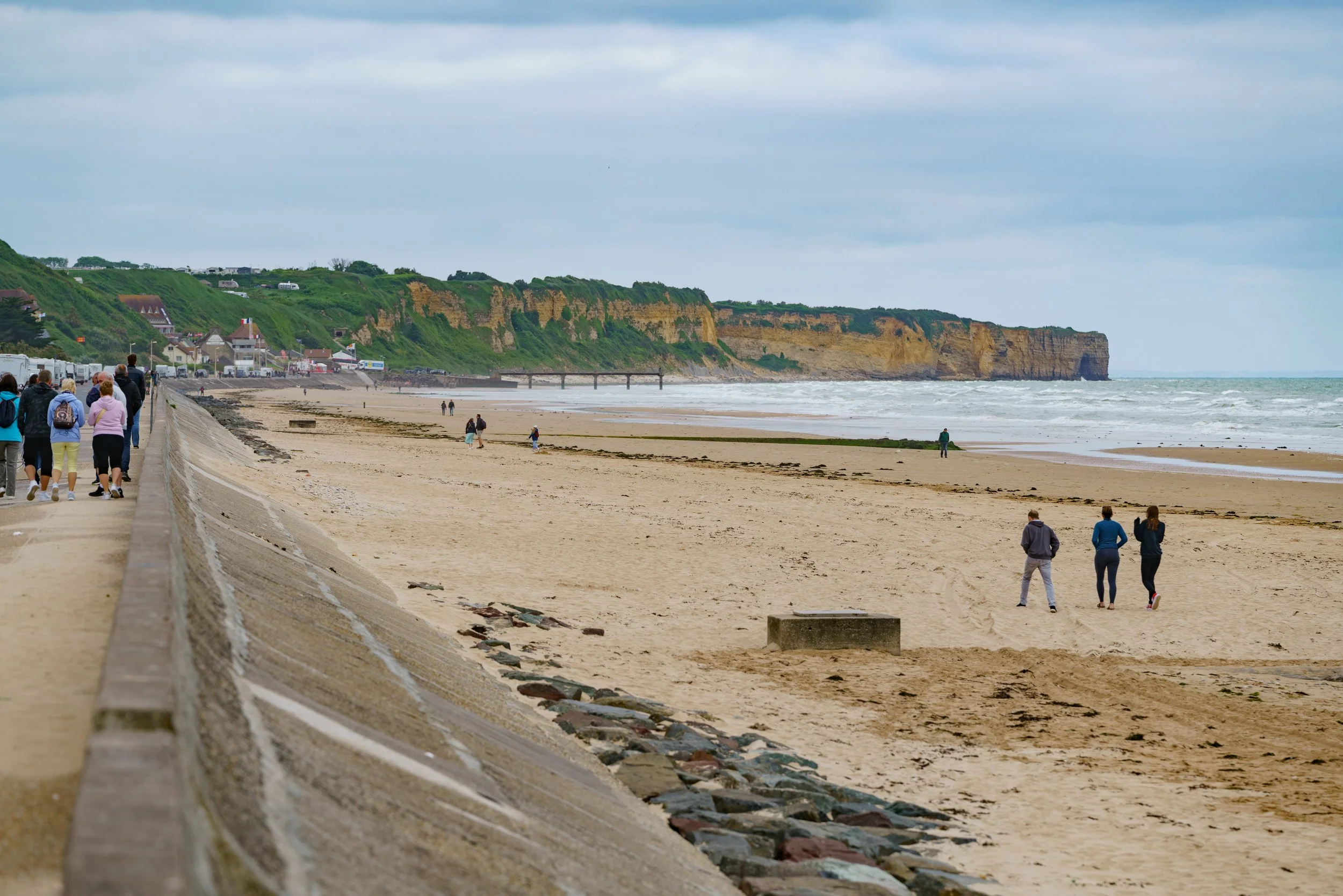 People walking along a beach promenade near sandstone cliffs, with some visitors on the sandy beach and others walking on the walkway.