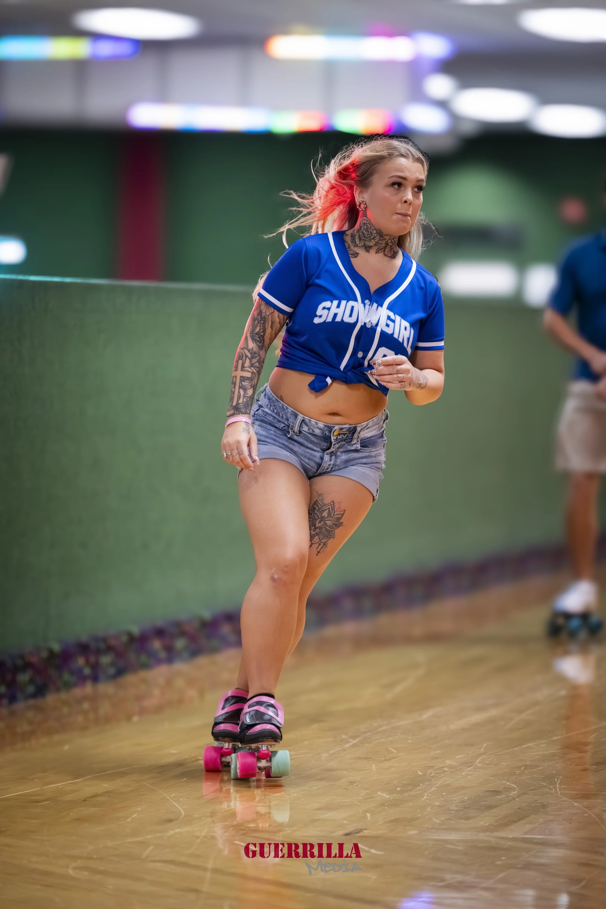 A woman skating indoors with a blue sports jersey, denim shorts, and tattoos visible on her arm and thigh.