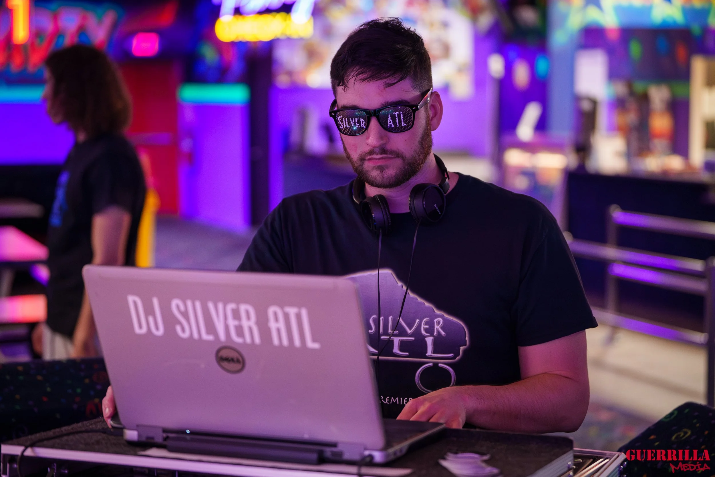 A man wearing sunglasses, headphones, and a black T-shirt with 'Silver ATL' on it, working as a DJ at a nightclub with colorful neon lights in the background.