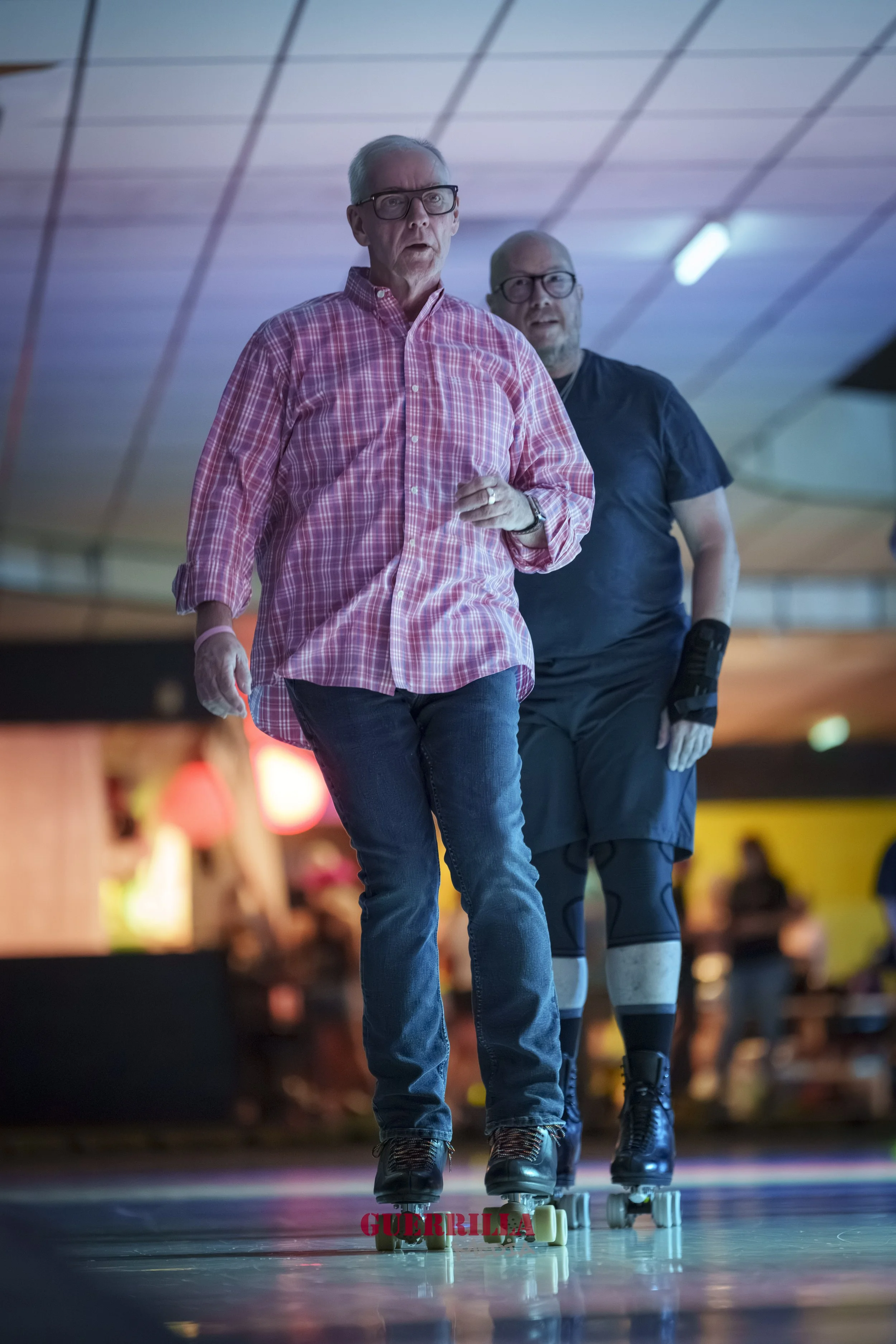 Two older men roller skating indoors, one wearing a pink plaid shirt and jeans, the other in black t-shirt and shorts, with a wrist brace, on a colorful, illuminated skating rink.