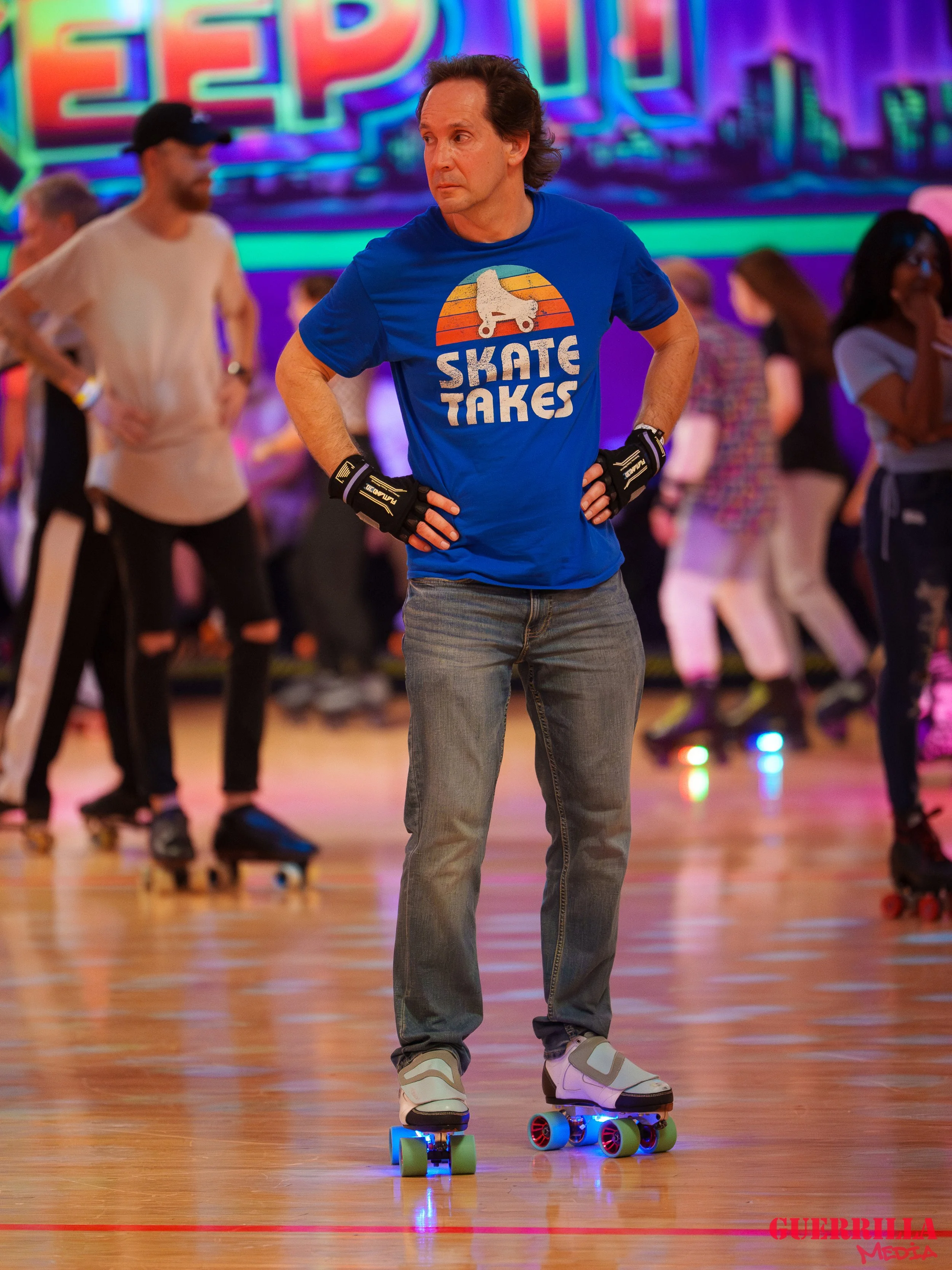 A man wearing a blue T-shirt with a roller skate graphic and the words "Skate Takes" is standing on roller skates at a roller rink. He has his hands on his hips and is surrounded by other skaters in the background.