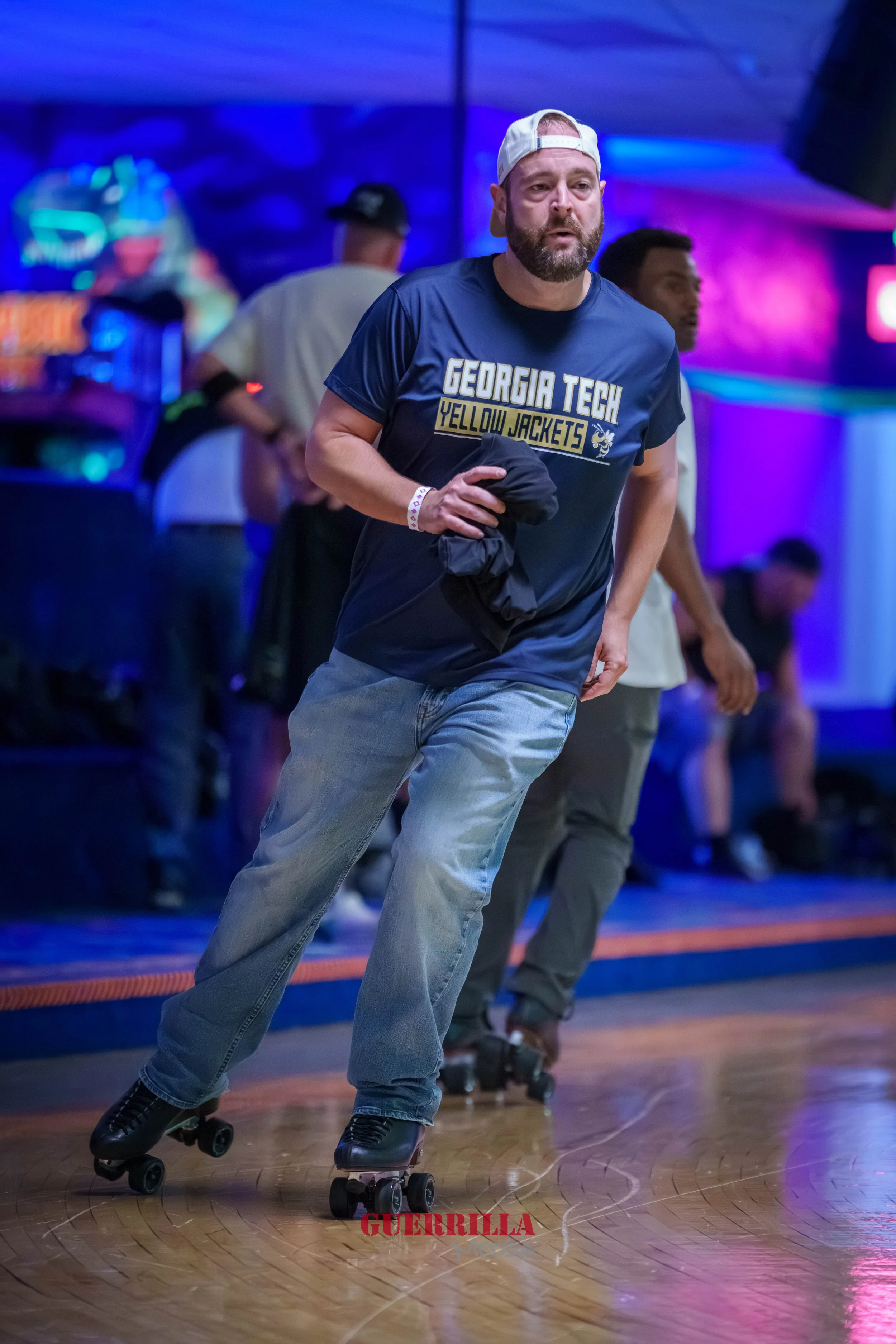 A man with a beard and white cap skating at an indoor roller rink, wearing a navy blue Georgia Tech Yellow Jackets t-shirt, jeans, and holding a black jacket, with other skaters and colorful neon lights in the background.