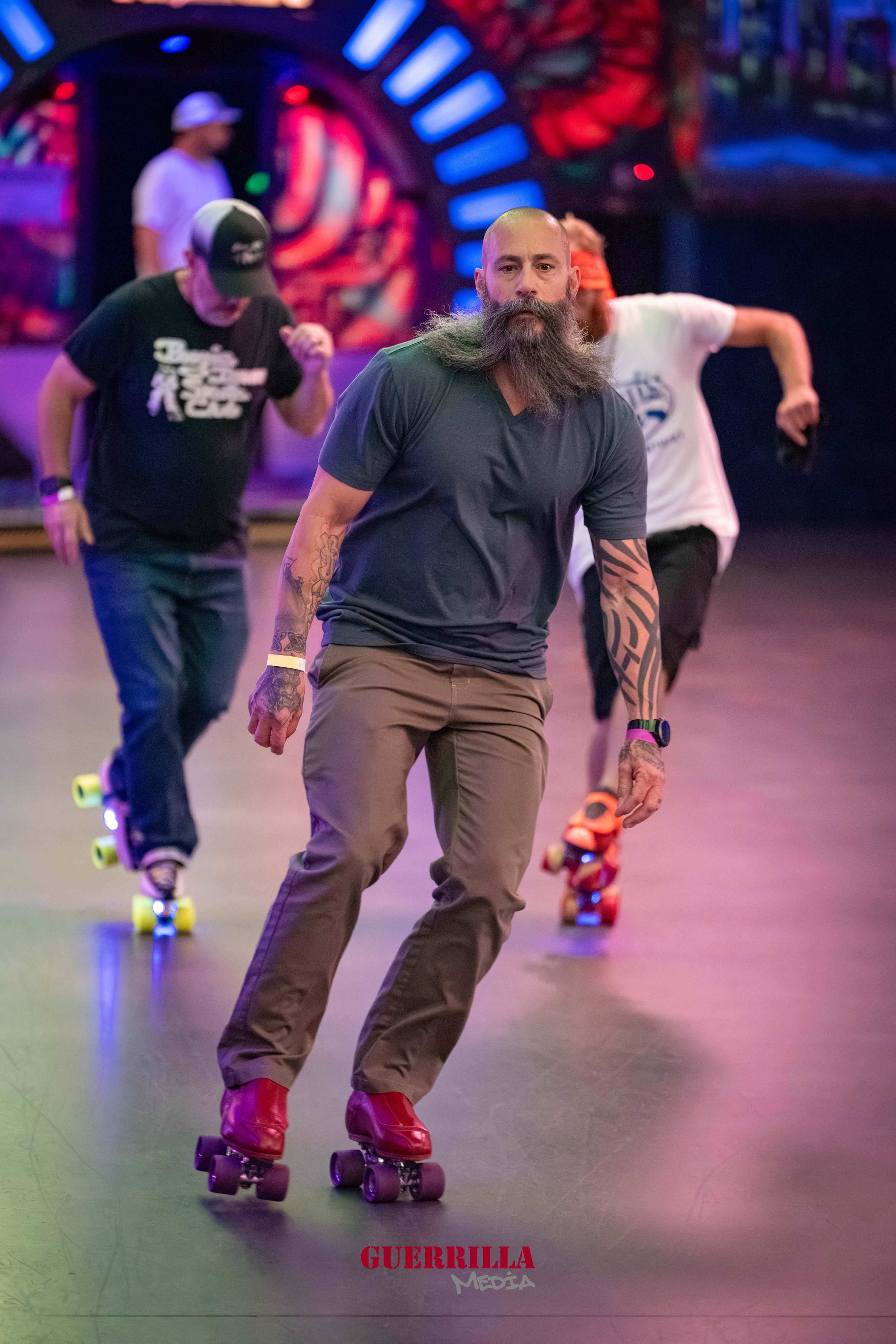 A bearded man with tattoos skating indoors with others in the background at an entertainment venue with colorful lights.