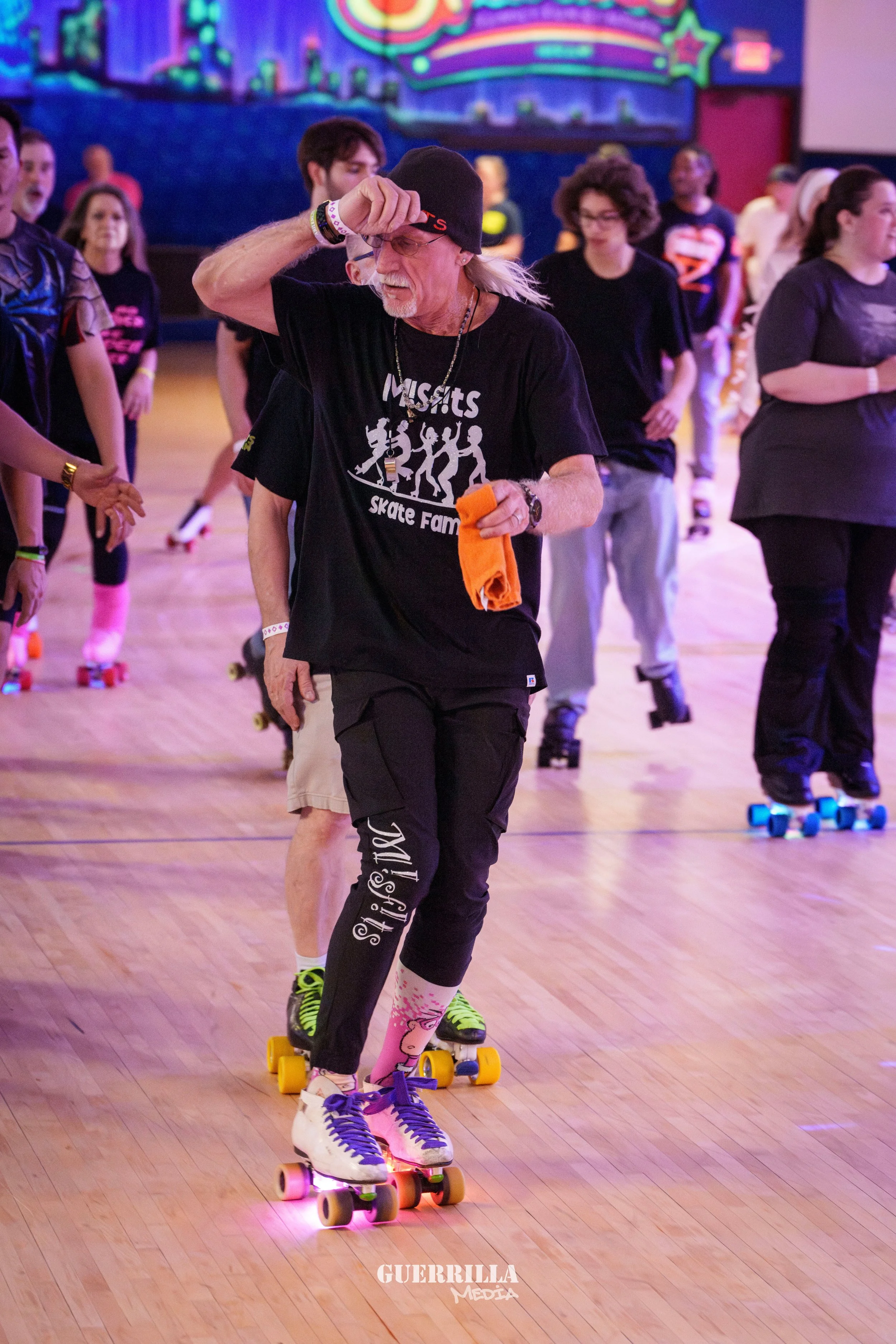 Older man skating on roller skate with colorful wheels, wearing a black beanie, black t-shirt, black pants, purple and white shoes, holding an orange cloth, at a roller skating rink with other skaters and a neon-lit background.