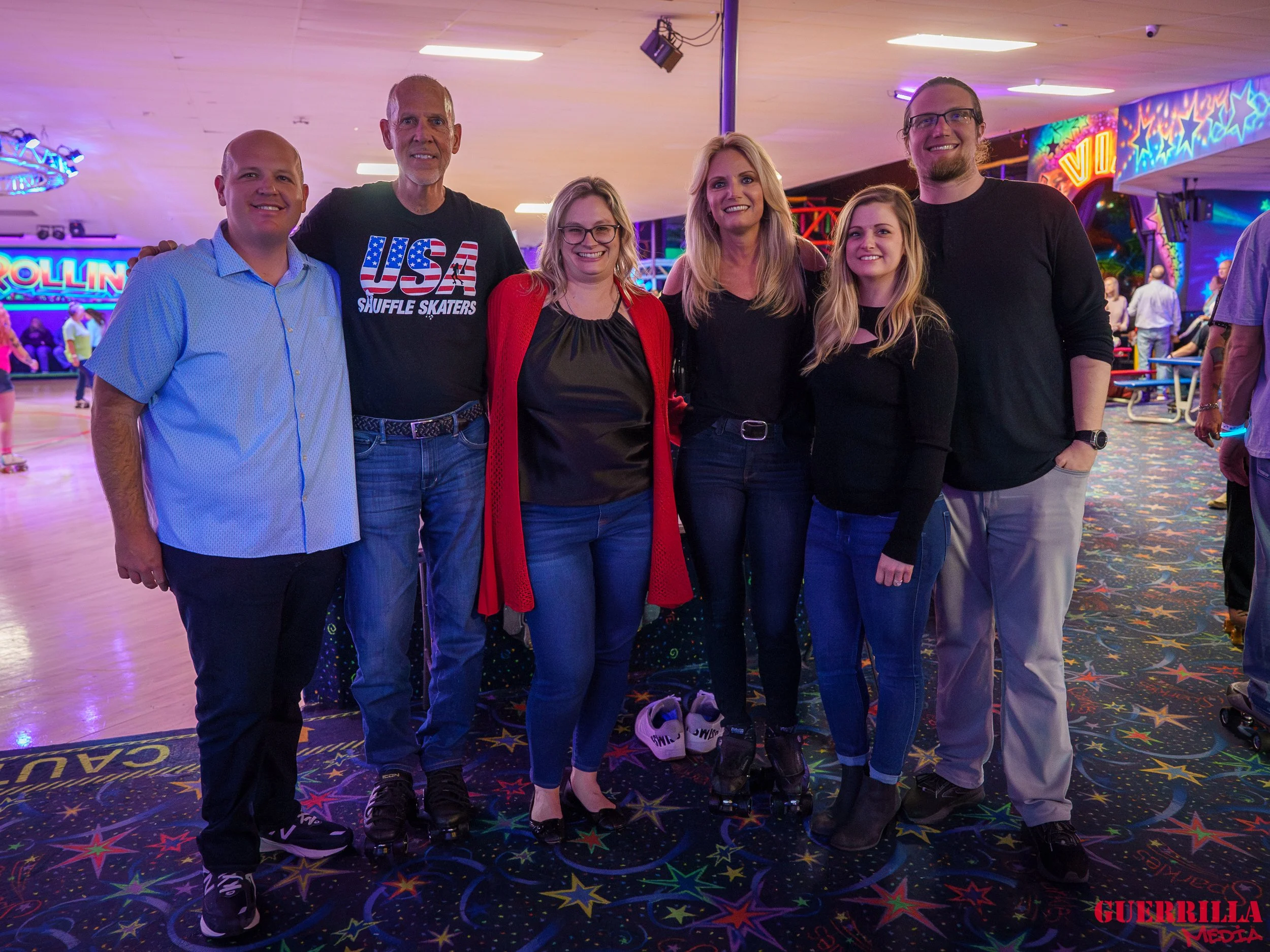 A group of seven people standing together at an indoor roller skating rink, smiling for a photo. The background features neon signs and colorful lights.