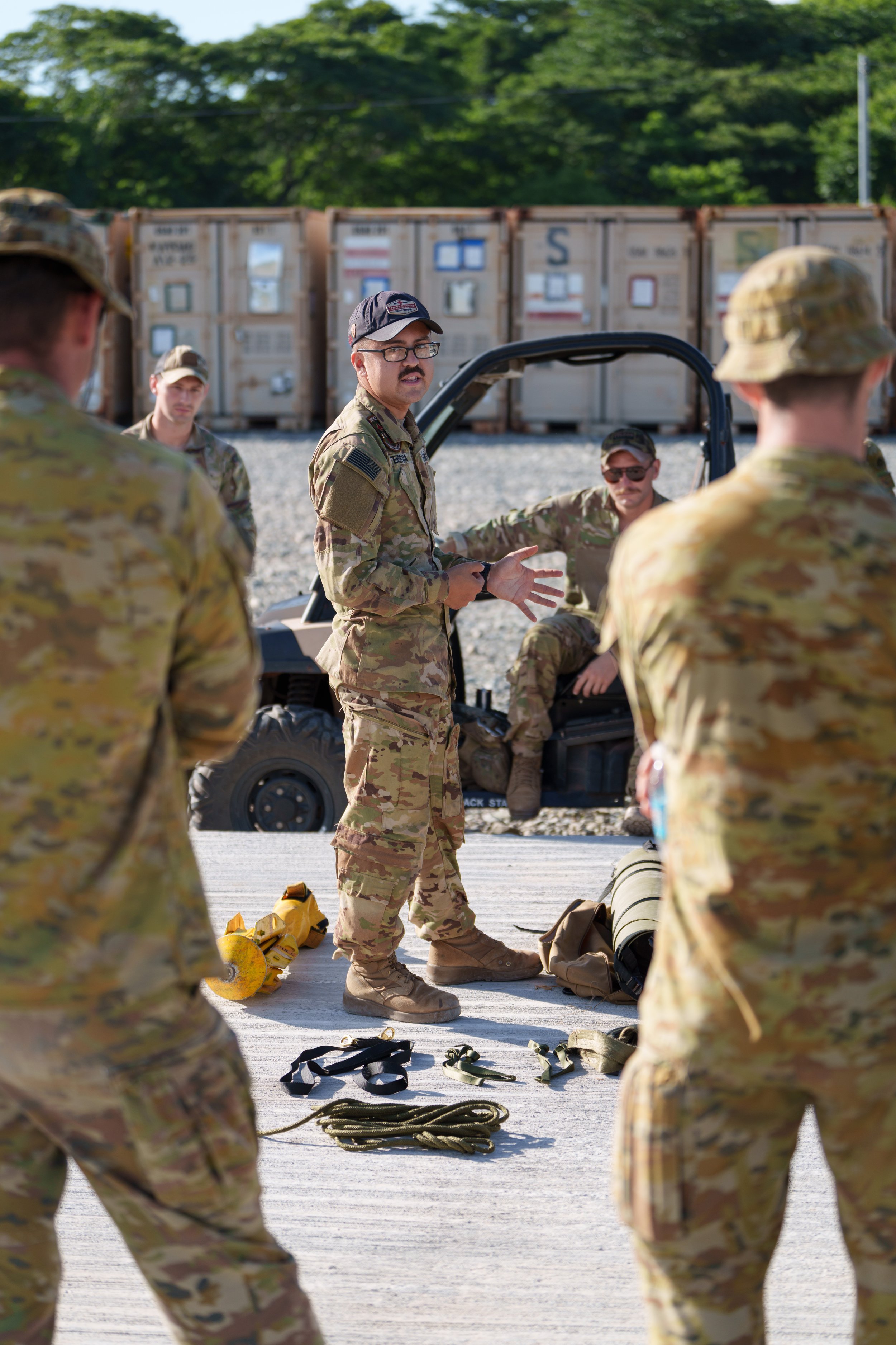 A military instructor in camouflage uniform and glasses demonstrating equipment to a group of soldiers in camouflage uniforms outdoors, with gear and a utility vehicle nearby.