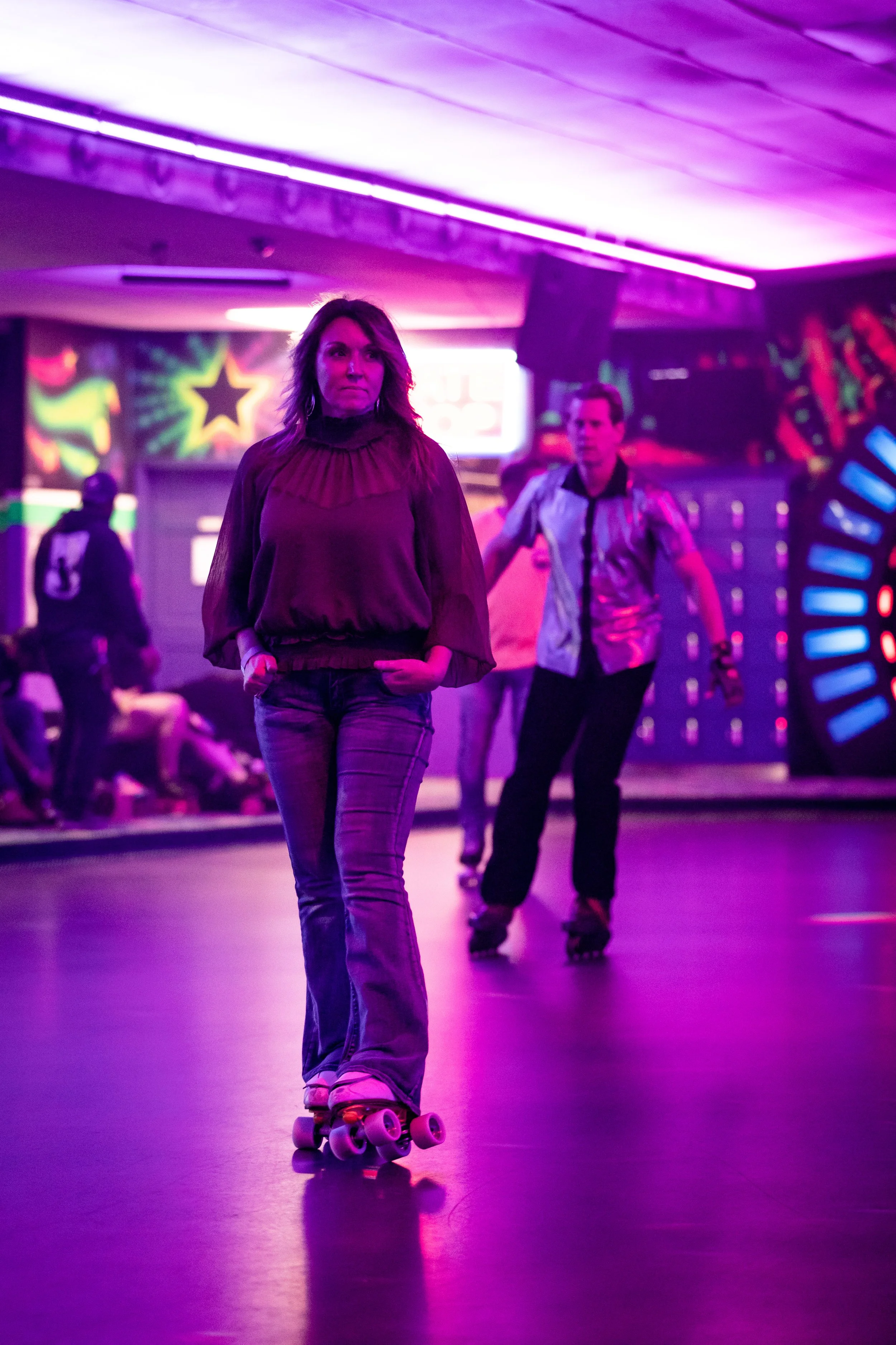 People roller skating in a neon-lit arcade or roller rink with vibrant purple and pink lights, arcade games in the background, and a woman in the foreground with her hands in her pockets.