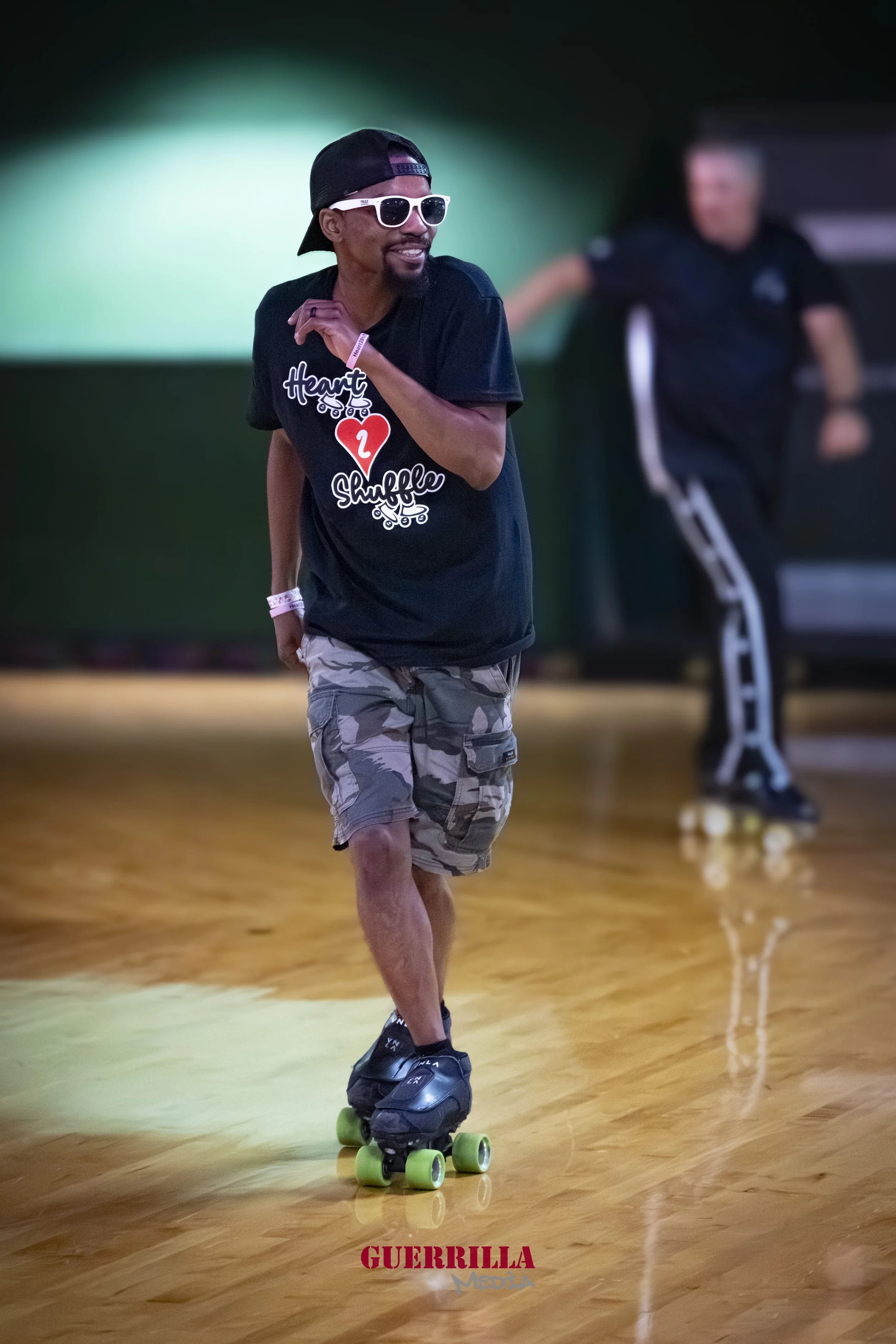 Man roller skating indoors, wearing sunglasses, a baseball cap, a black t-shirt with 'Heart Shuffle 2' graphic, and camouflage shorts, with other skaters in the background.