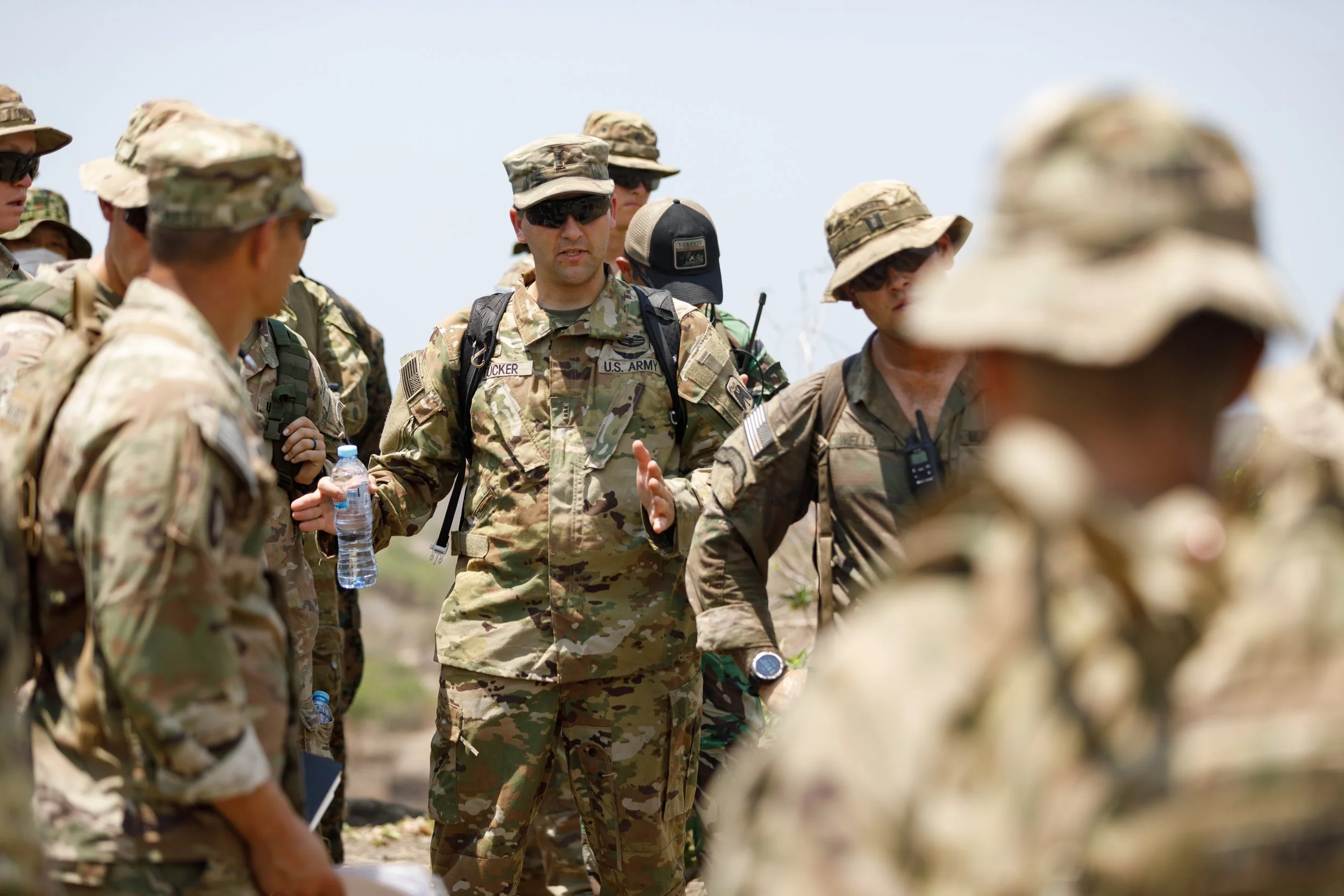 A group of soldiers in camouflage uniforms gathered outdoors, listening to a person who appears to be giving instructions or a briefing.