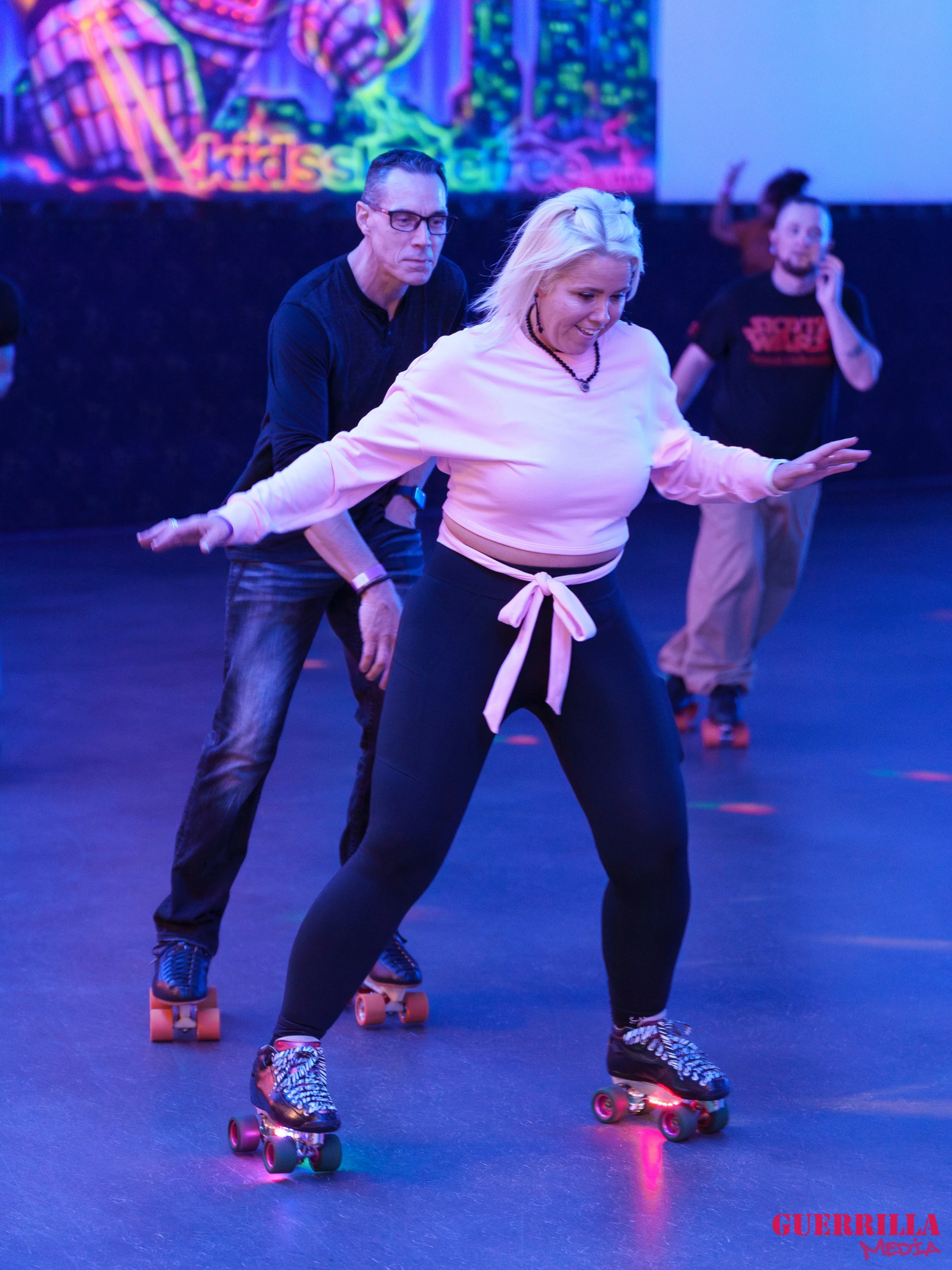 A group of people roller skating in an indoor rink with neon lights. One woman is in the foreground, balancing on roller skates, with a man behind her guiding her. Other skaters are in the background, enjoying the activity.