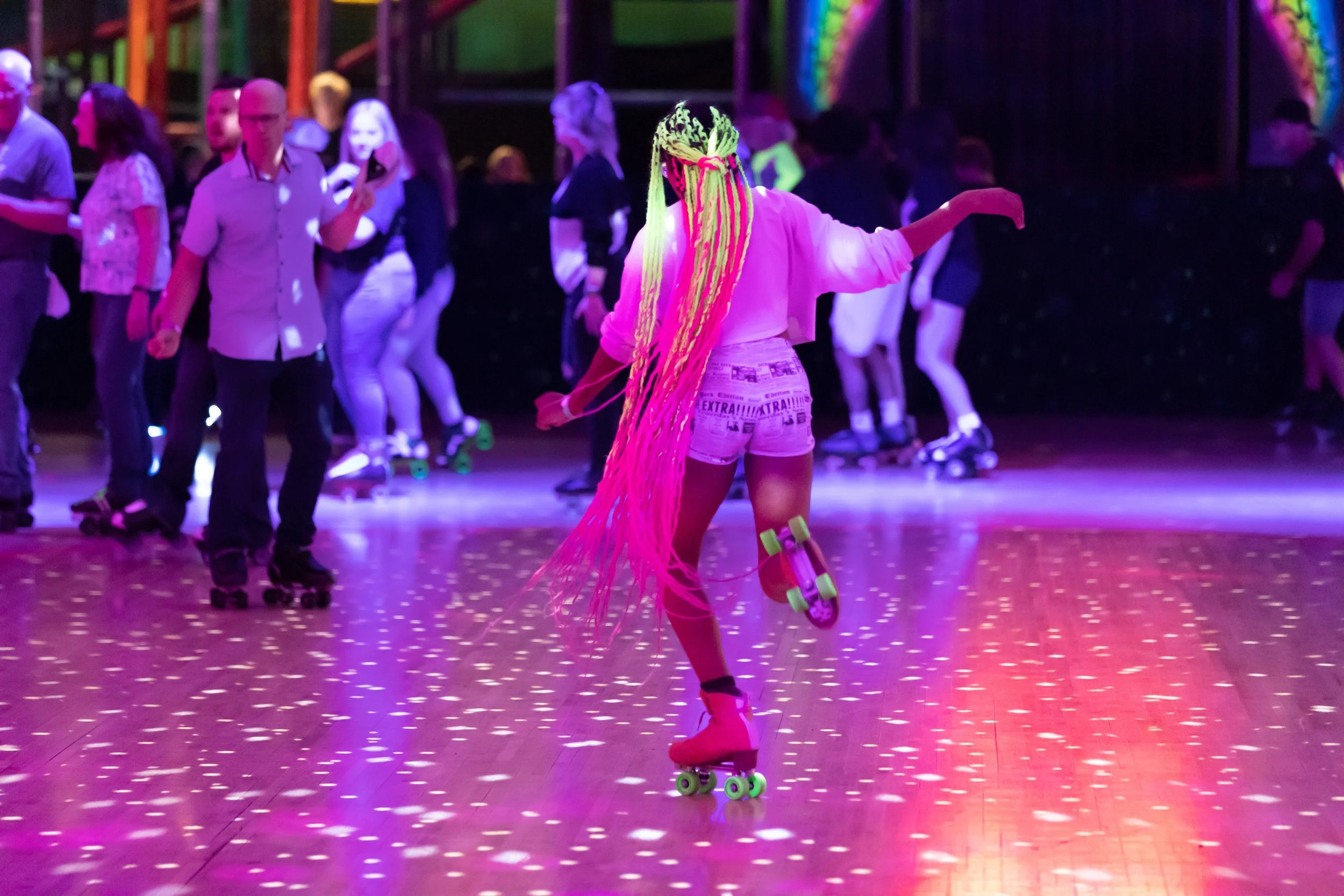 A woman roller skating on a dance floor with colorful lights, wearing neon orange skates, pink hair extensions, and a casual outfit, surrounded by other skaters at a roller disco or club.