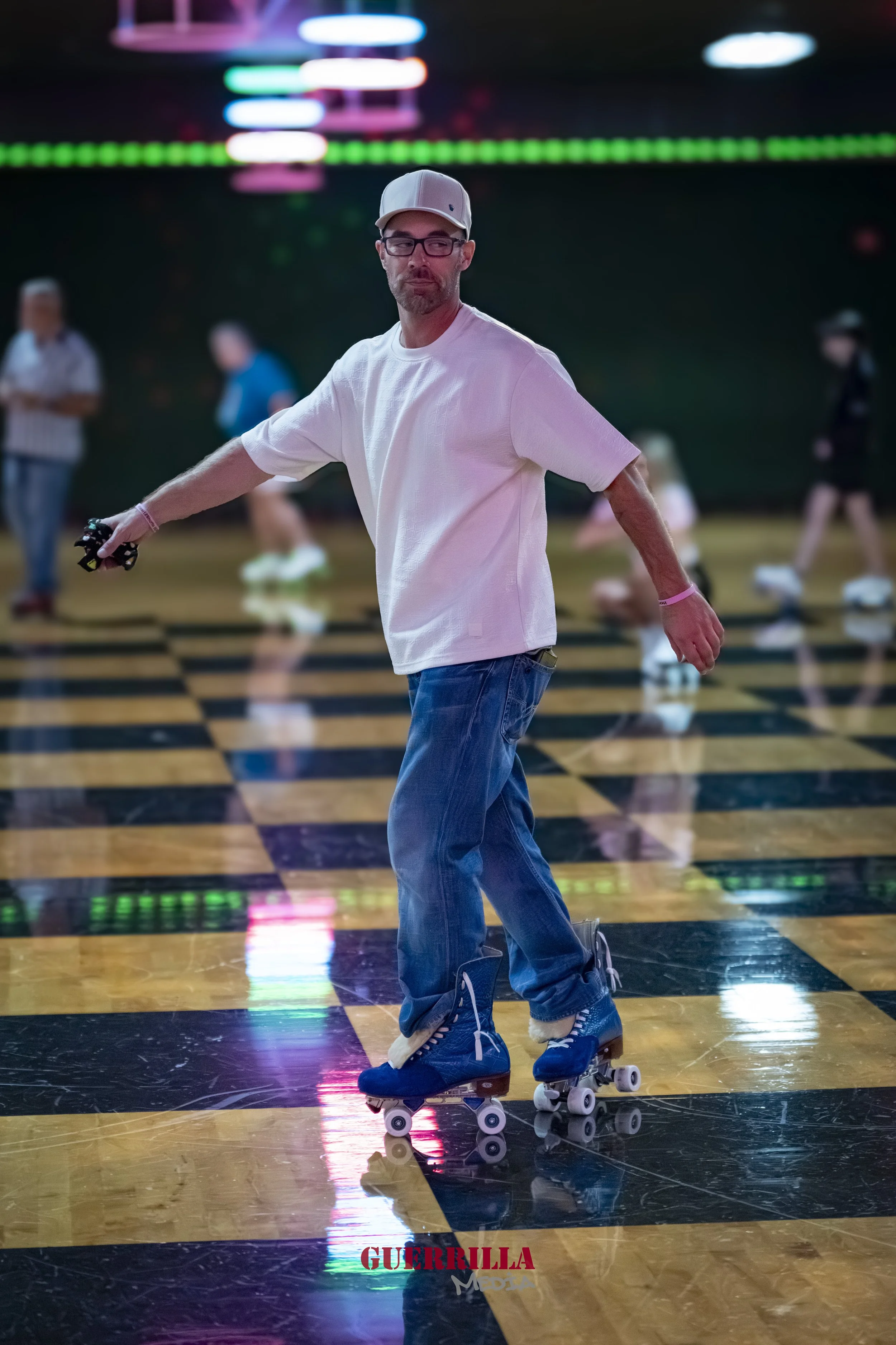 A man wearing glasses, a gray baseball cap, a white t-shirt, and jeans roller skating on a checkered black and yellow floor in a roller rink with neon lights and other skaters in the background.