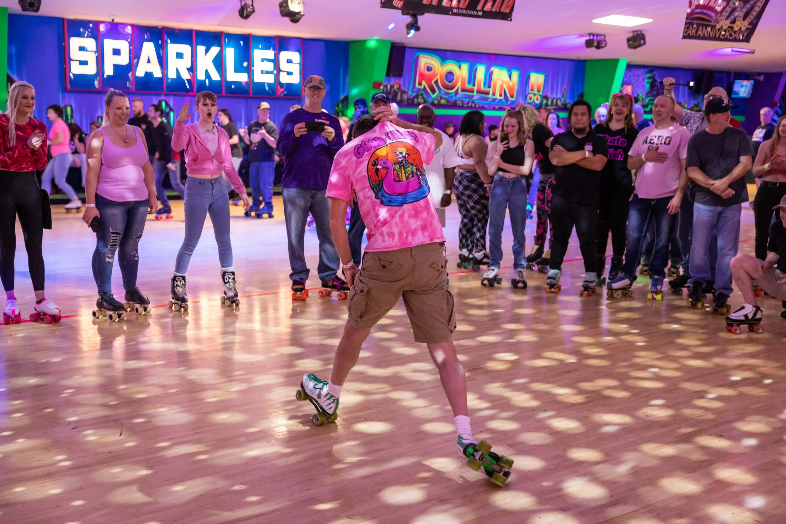 People roller skating at an indoor roller rink with colorful neon signs that say 'Sparkles' and 'Rollin'. The center person is wearing a pink tie-dye shirt and beige shorts, skating in front of a group watching and taking photos.
