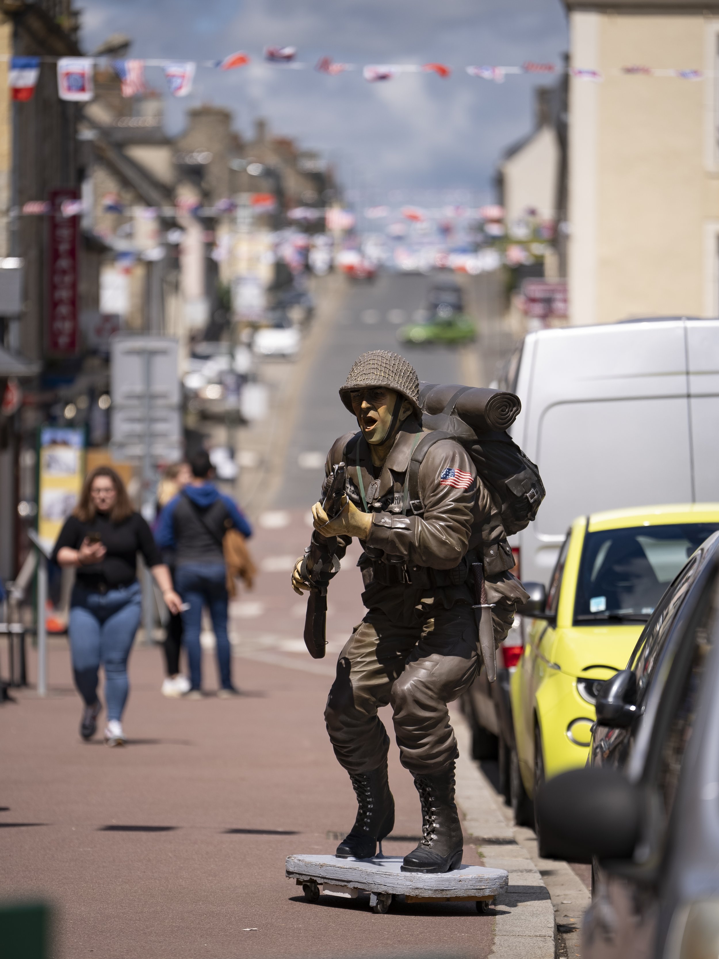 Street performer dressed as a soldier, painted face, wearing a helmet, standing on a skateboard, in an urban street with pedestrians and parked cars.