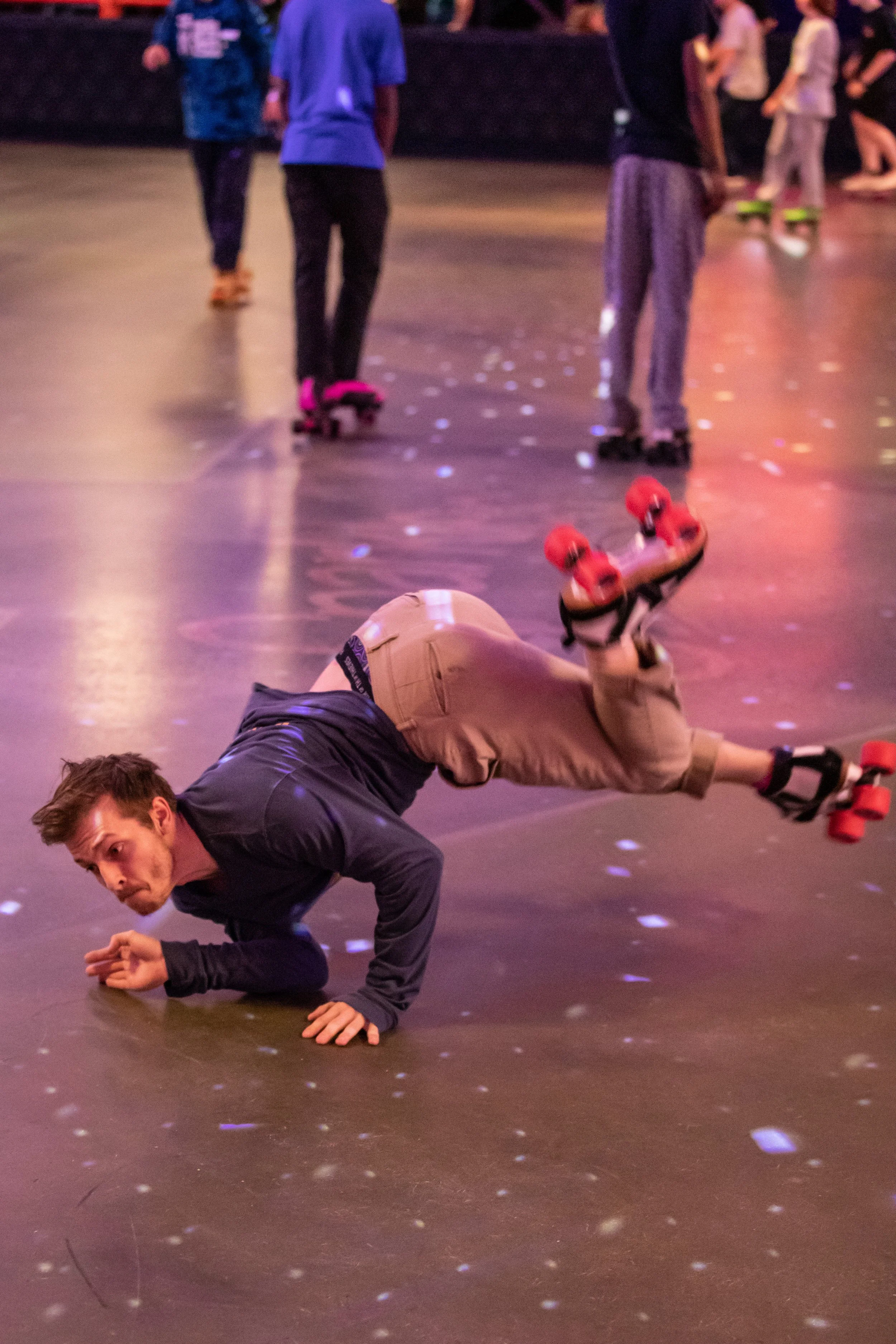 A man falls and is on the floor in a roller skating rink, with his body upside down and one arm supporting him. Other people are skateboarding in the background.