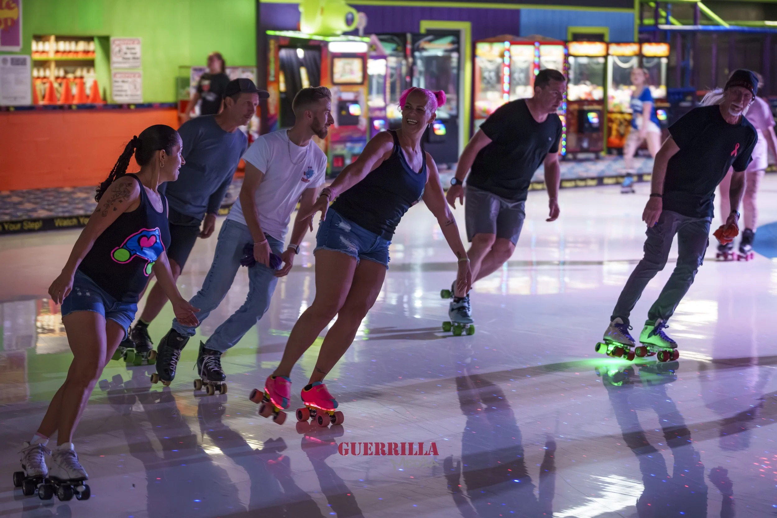 Group of six people roller skating in an indoor arcade or roller rink, with colorful arcade games in the background and a reflective floor.