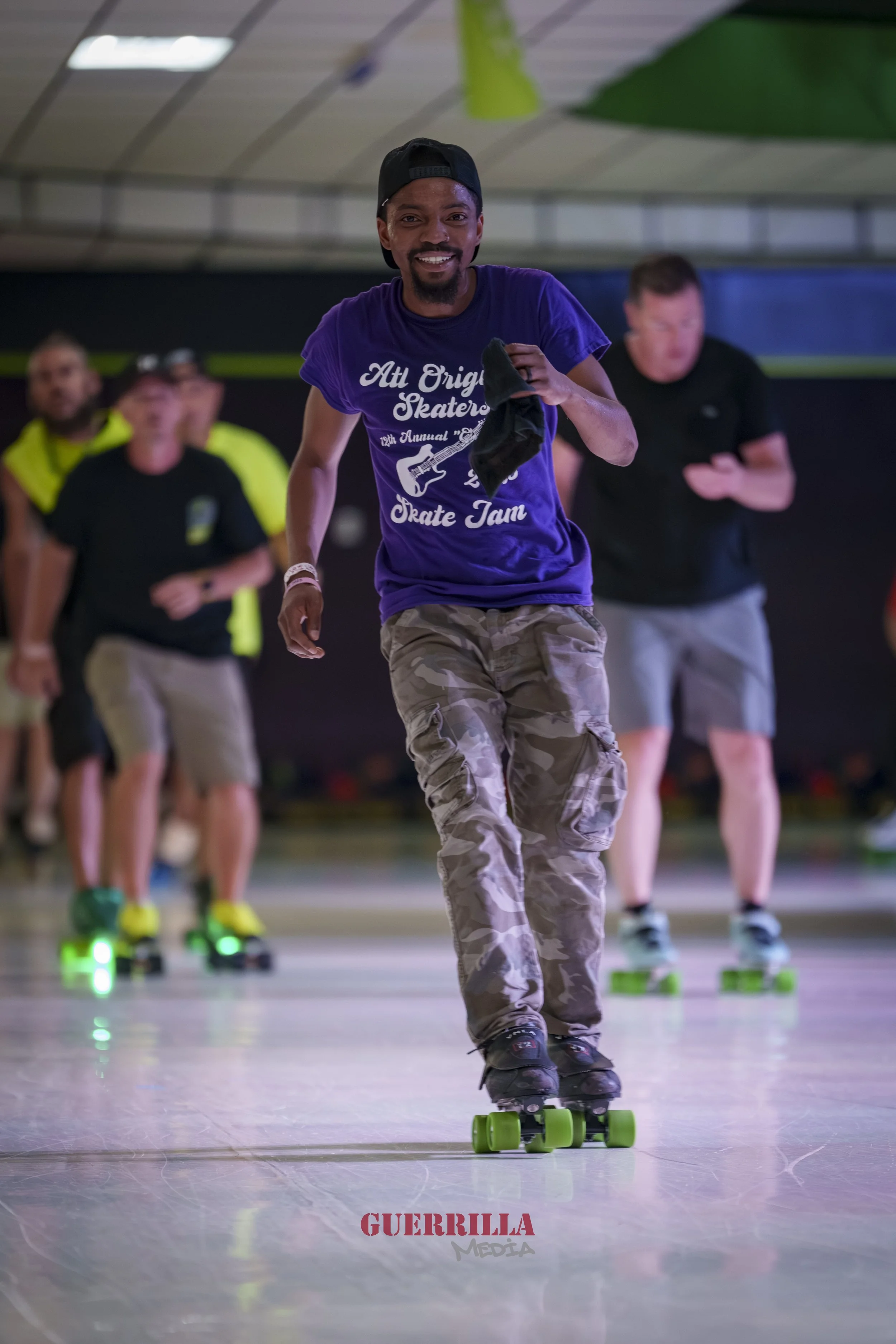 A man smiling and roller skating on an indoor rink, holding a black cloth in his right hand, with other people roller skating in the background.