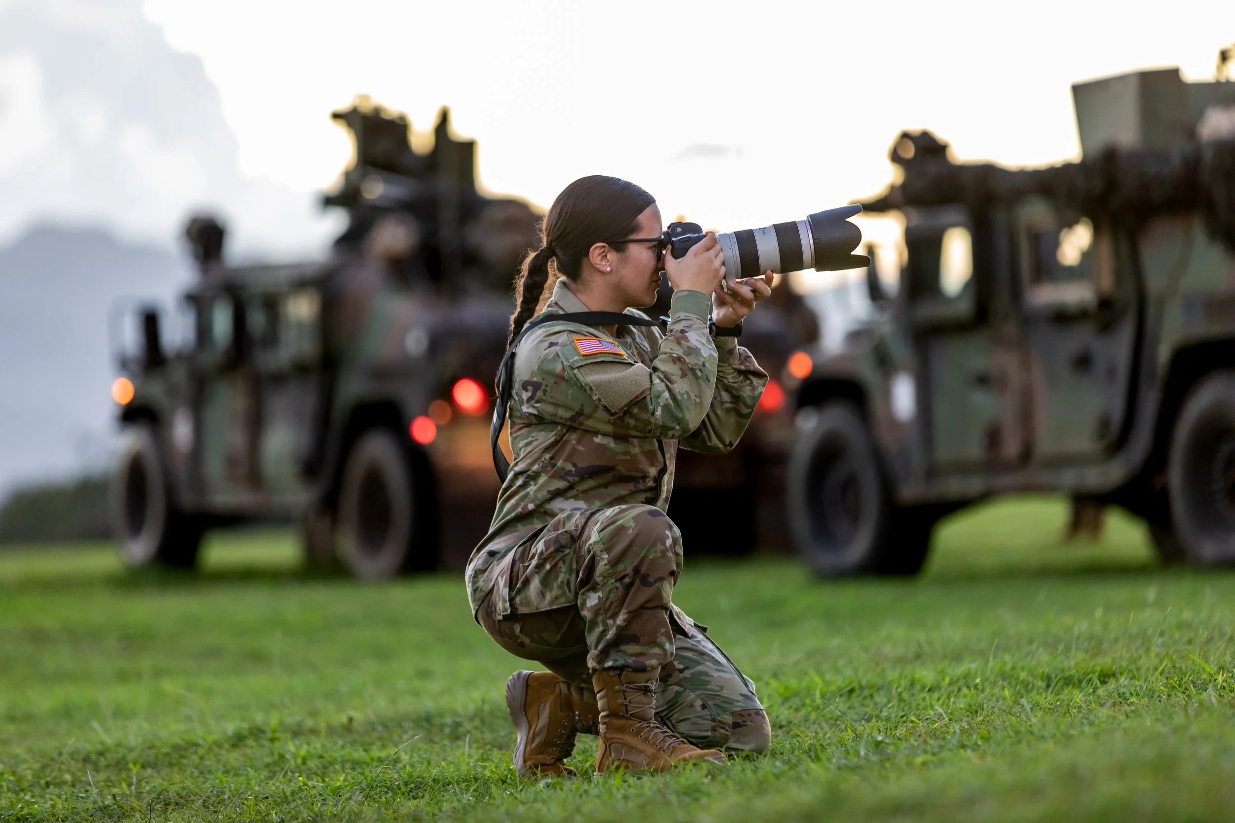 A woman in military uniform kneeling on the grass and photographing with a large telephoto camera lens, with military vehicles in the background.
