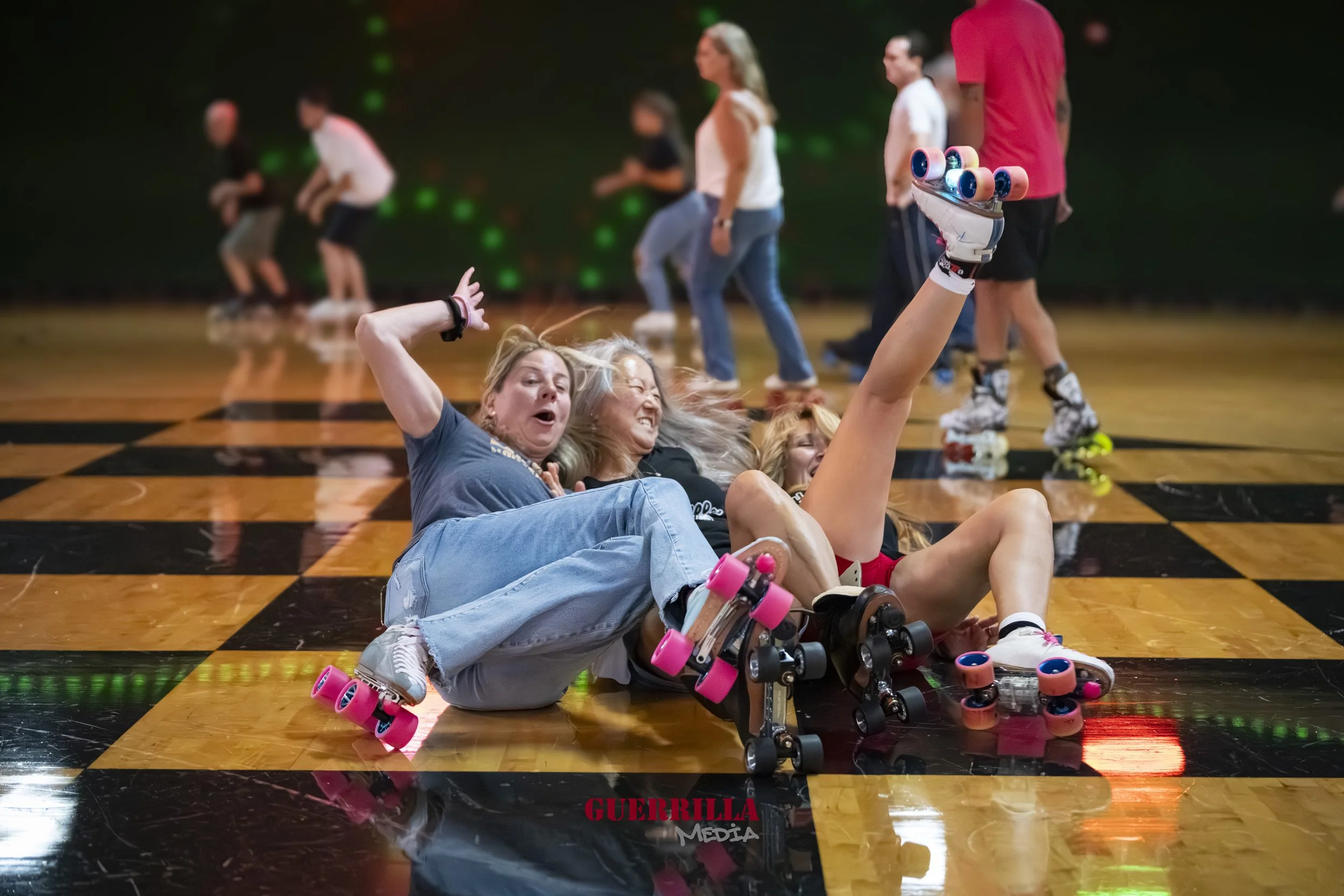 Two women wearing roller skates falling and laughing on a checkered floor at a roller skating rink while other skaters are in the background.