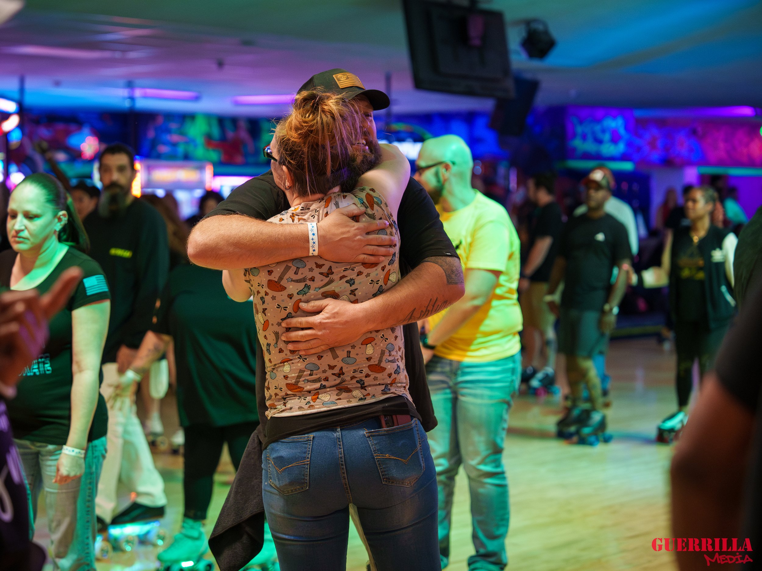 Two people hugging on a dance floor at a lively social event with colorful neon lighting and other attendees in the background.