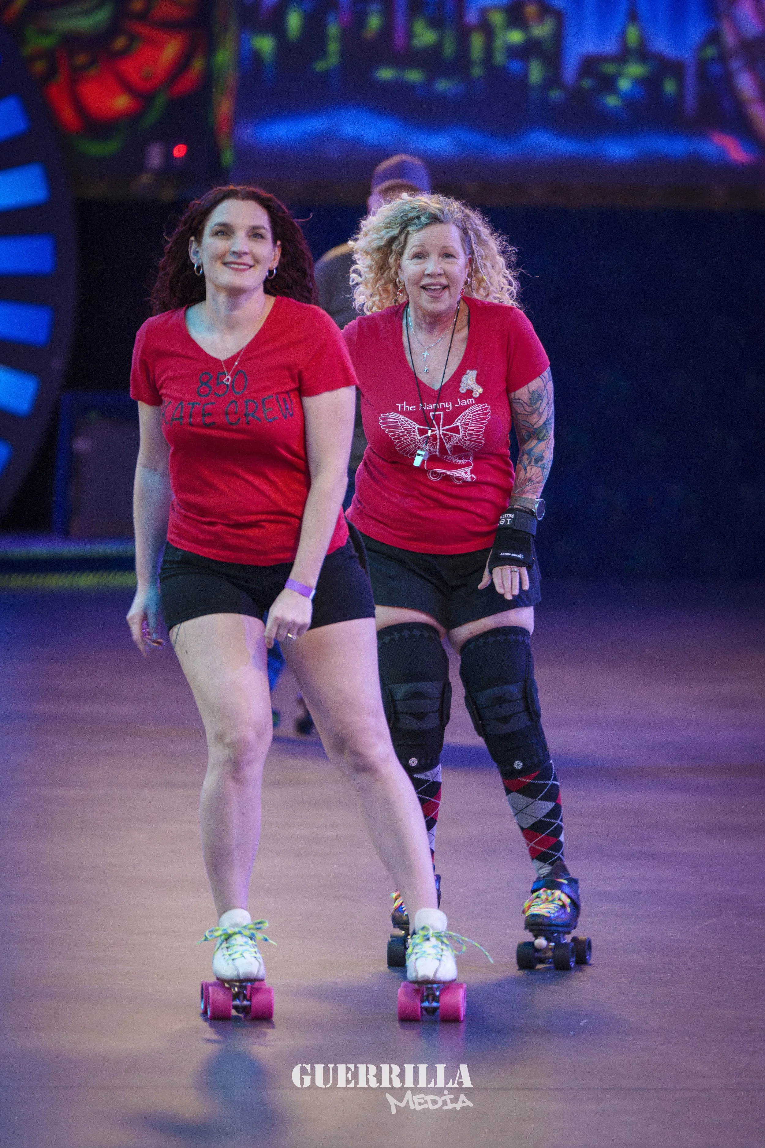 Two women roller skating indoors, wearing red shirts, with a colorful background and a logo that says 'GUERRILLA MEDIA' at the bottom.