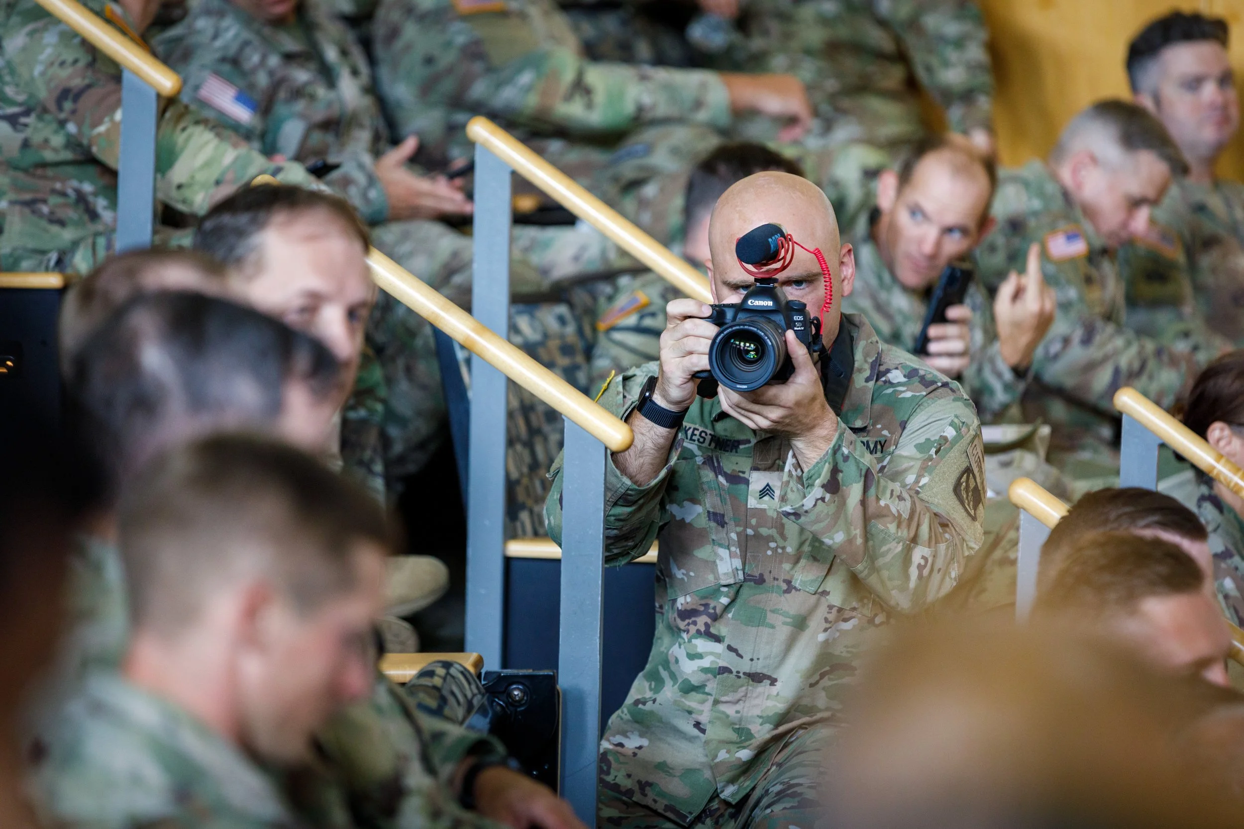 A group of soldiers in camouflage uniforms seated in an auditorium. One soldier in the center is taking a photo with a professional camera.