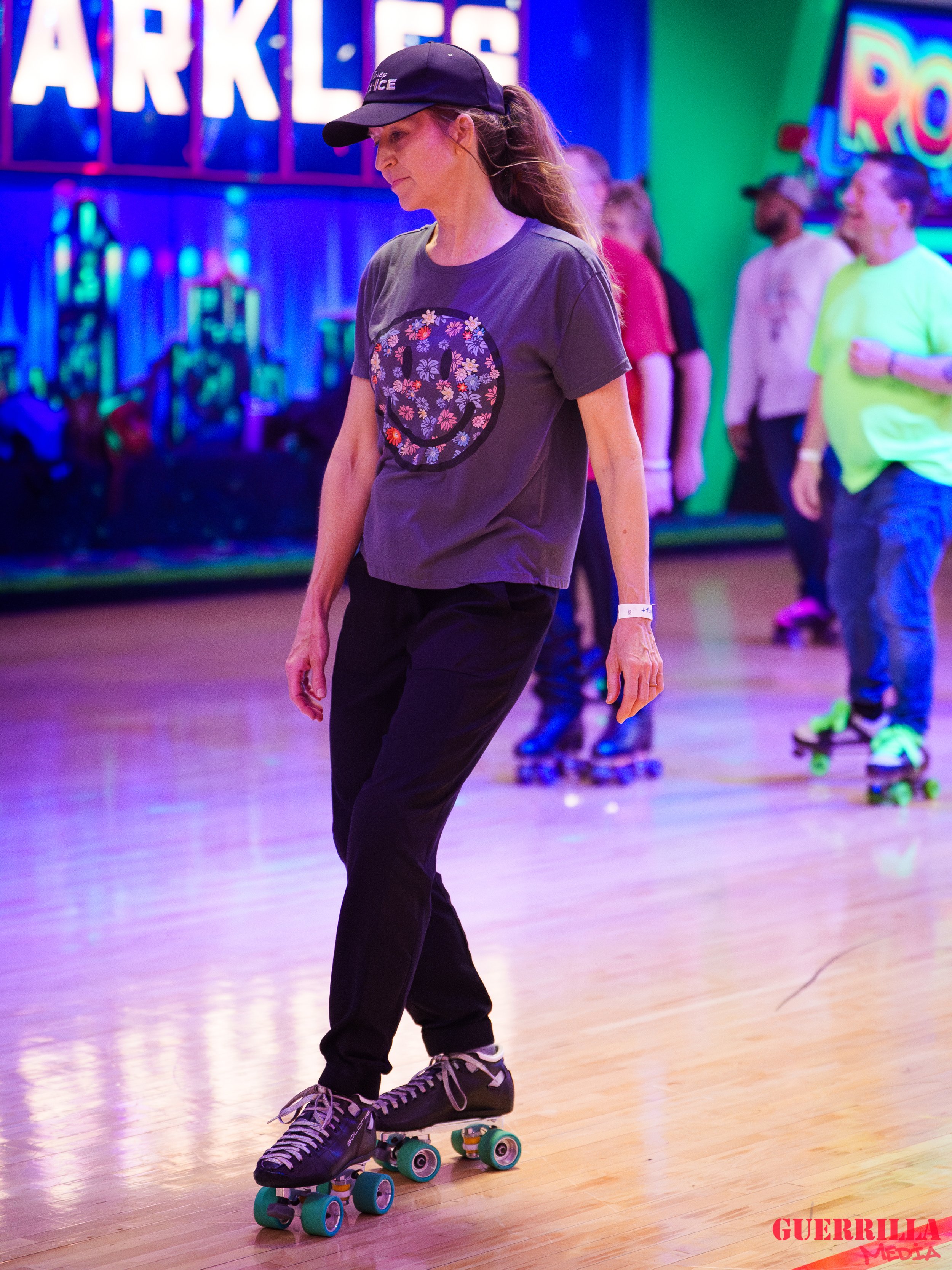 A woman roller skating indoors, wearing a black cap, gray T-shirt with a smiley face made of flowers, black pants, and roller skates with black laces and teal wheels. Other people are in the background also roller skating in a colorful arcade setting