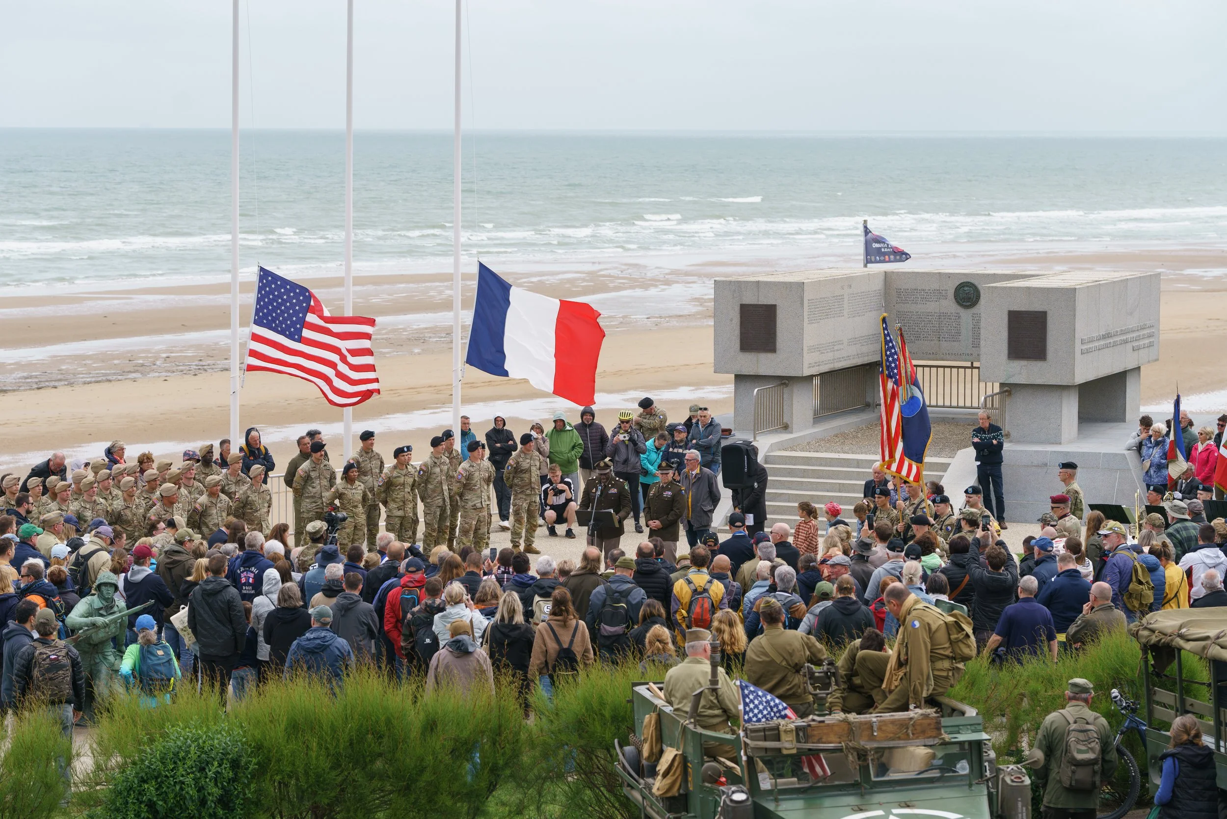 A large group of people attending a memorial event at a beach, with American and French flags flying, military personnel in uniform, and a monument in the background.