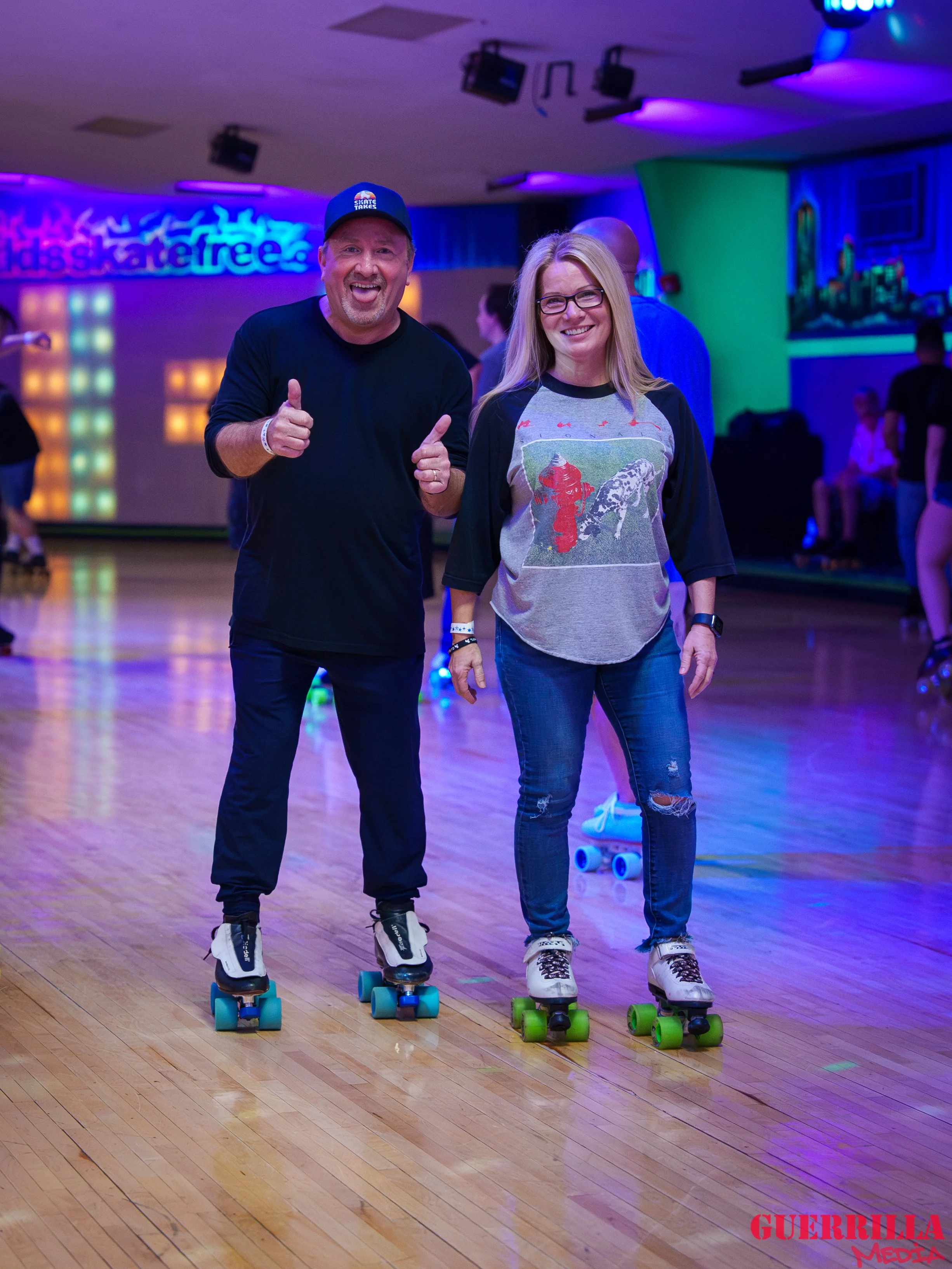 A man and a woman roller skating on a wooden floor at an indoor roller rink. The man is giving a thumbs up and wearing a cap, black shirt, and black pants. The woman is smiling, wearing glasses, a gray and black shirt, and ripped jeans. There are oth