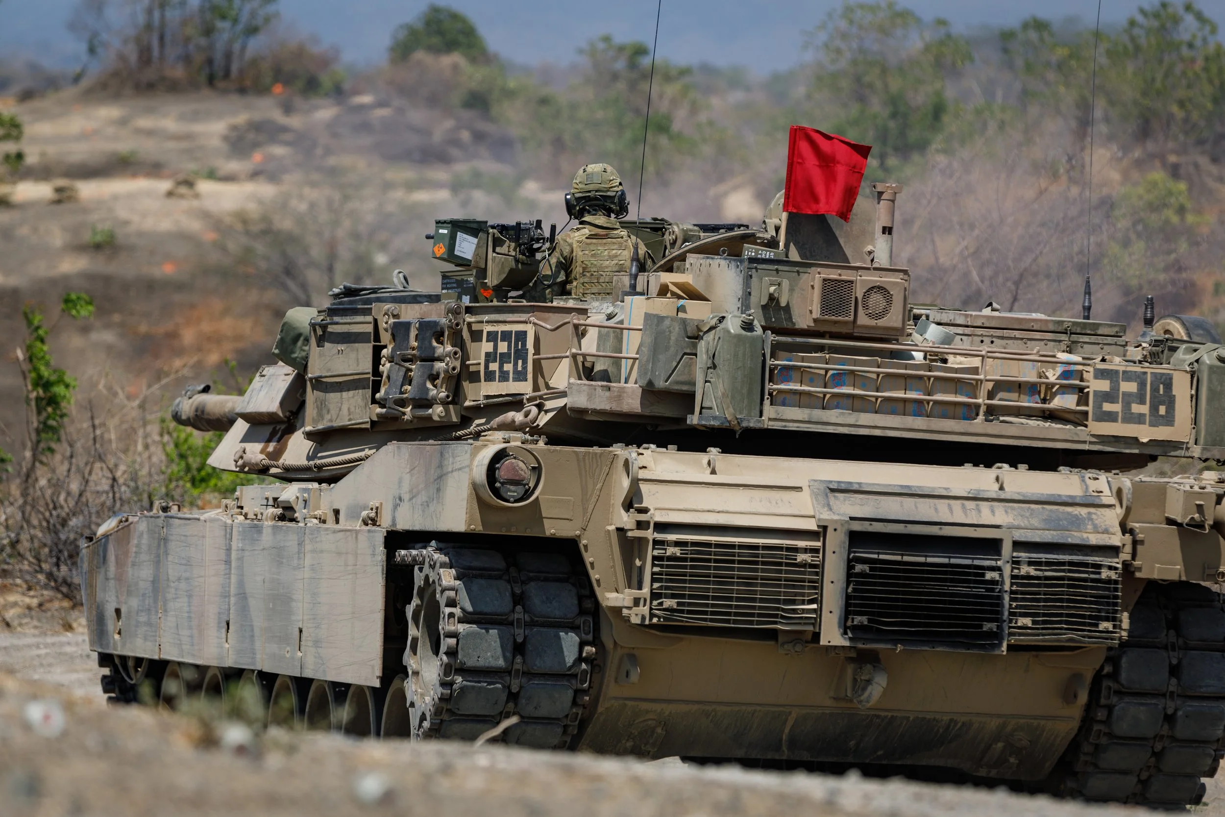 A military tank with a soldier operating its gun, moving across a rugged landscape with sparse vegetation and trees in the background.