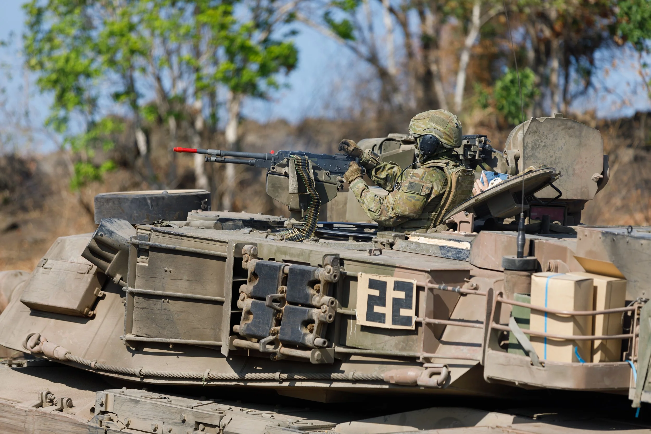 Soldier in camouflage uniform operating a mounted machine gun on a military tank, in an outdoor setting with trees in the background.